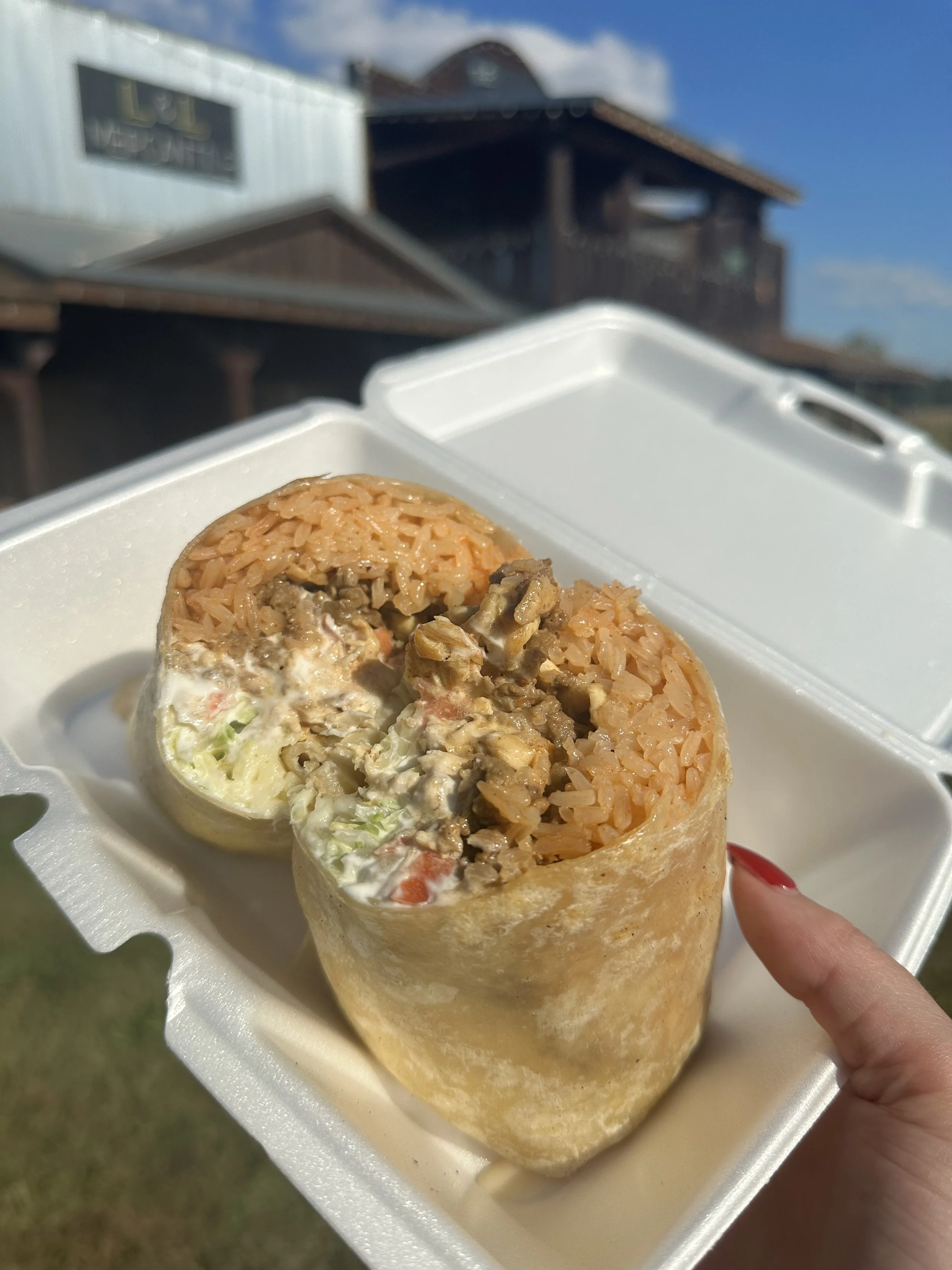 Close-up of a wrap sushi with rice, vegetables, and meat in a foam container. A person is holding the container with a thumb and finger, with a finger nail painted red. In the background, there are buildings and a blue sky.
