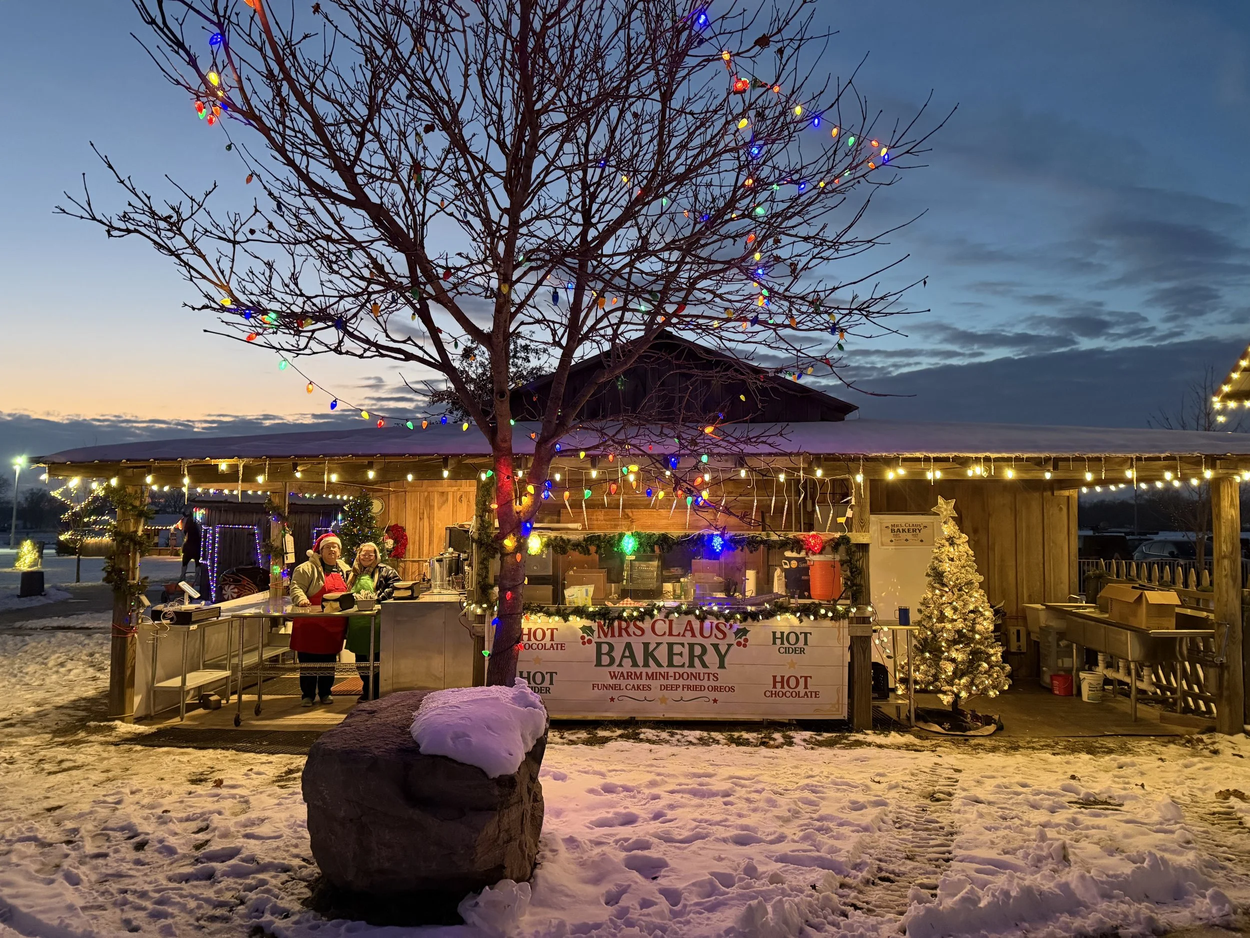 A snow-covered outdoor scene at dusk featuring a decorated tree with colorful Christmas lights, a wooden stall selling baked goods with a sign reading 'Mrs. Claus Bakery', and a large rock in the foreground with snow on top. There are people behind the stall, and the sky is twilight with clouds.