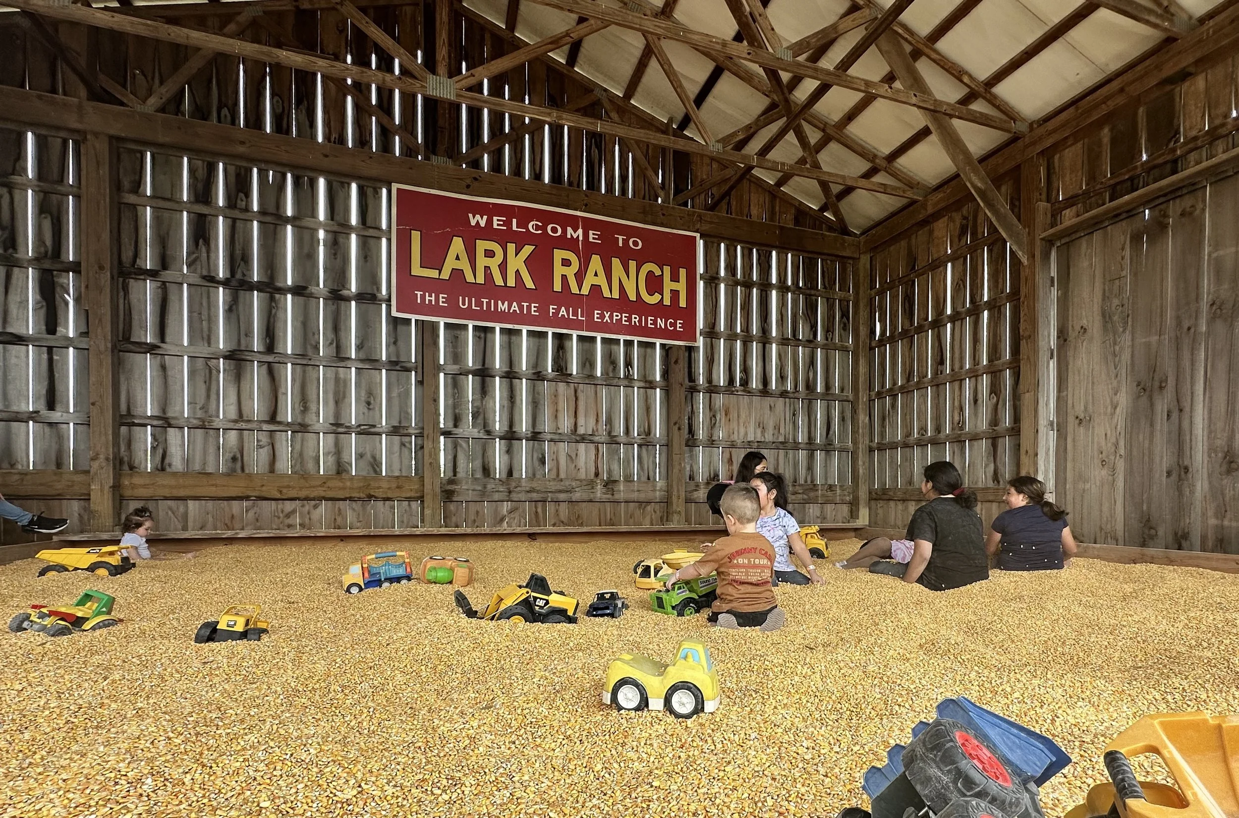 Children playing with toy trucks on a corn maze ground inside a barn, with a large wooden wall and a sign that says "Welcome to Lark Ranch, The Ultimate Fall Experience."