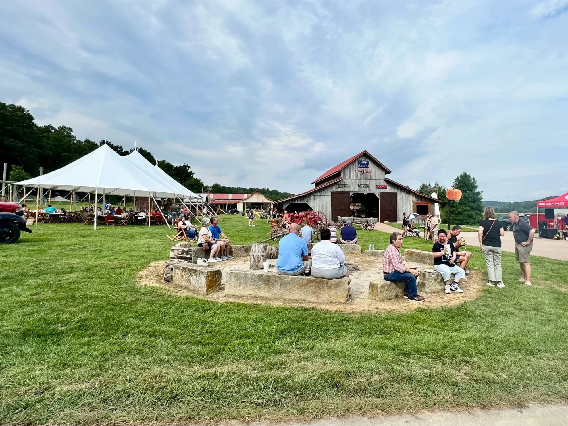 A rural outdoor event with a large white tent, a barn labeled 'Lark Ranch,' and people sitting on benches and walking on a green grassy area. The sky is partly cloudy and there are trees in the background.