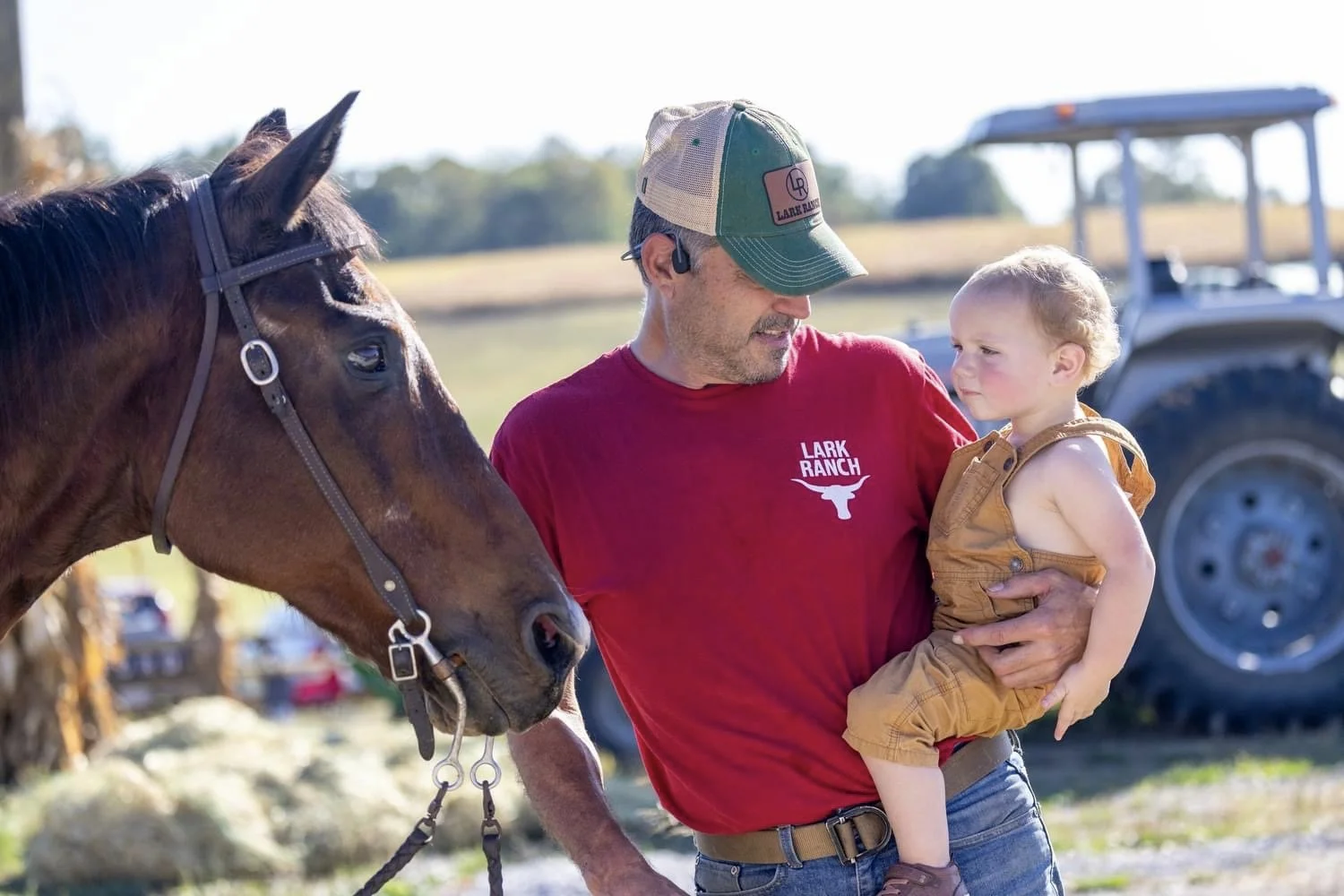 A man holding a young boy outdoors near a horse, with a tractor in the background.