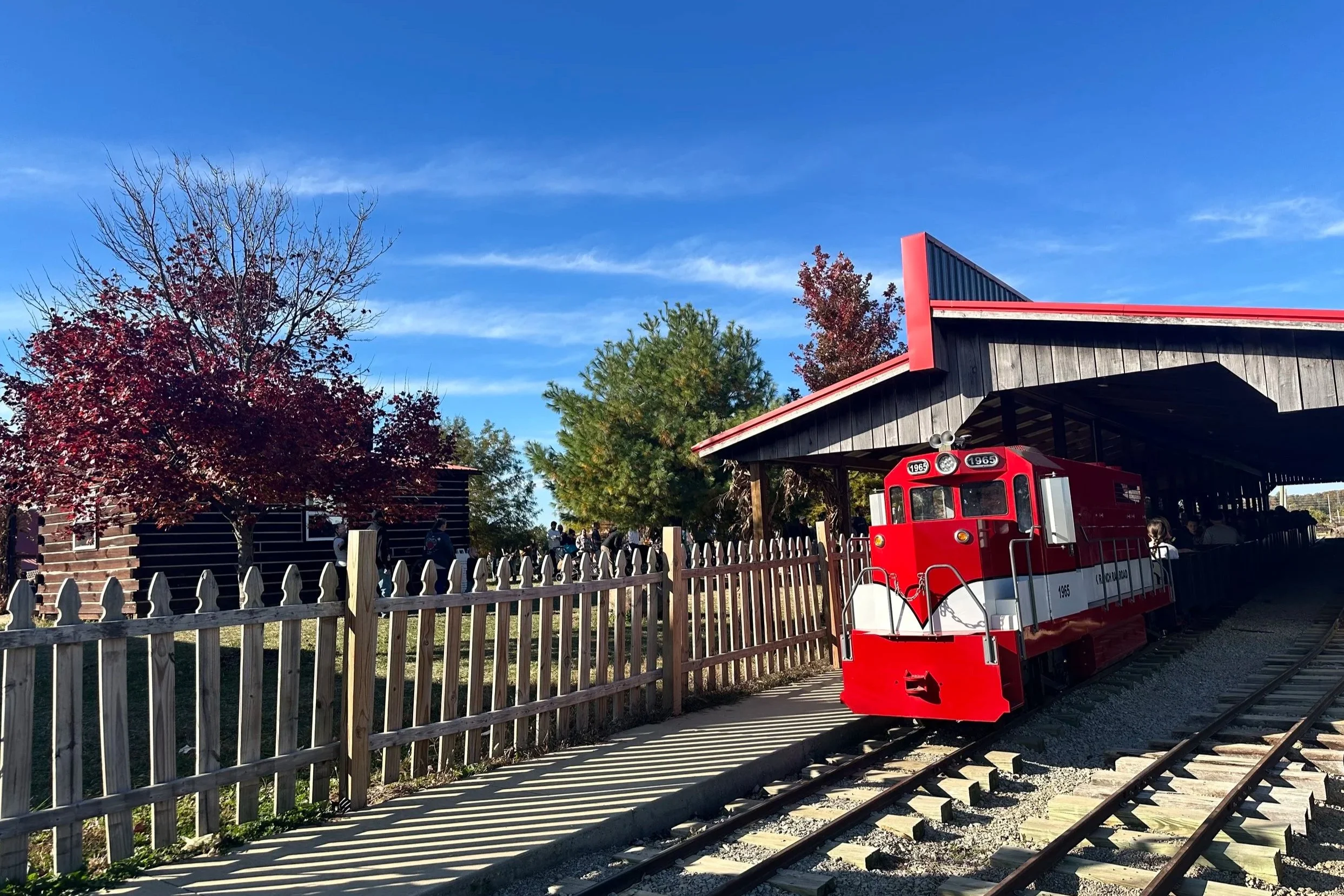 A small red train engine on a track outside a wooden station with autumn trees and a group of people.