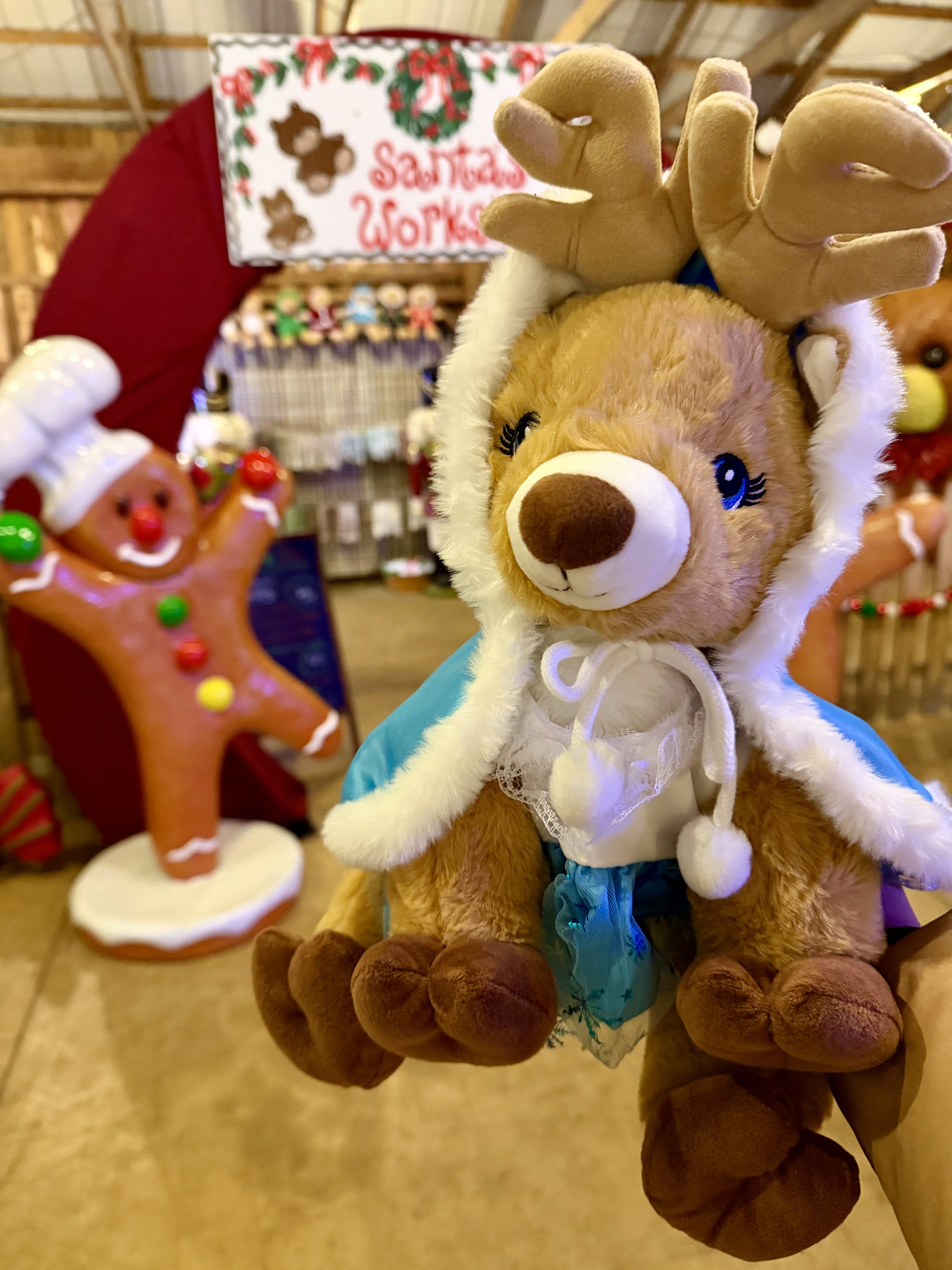 Close-up of a plush teddy bear wearing reindeer antlers and a white hooded cape. In the background, there is a gingerbread cookie decoration with colorful candies and a 'Santa's Workshop' sign with Christmas themed decorations.