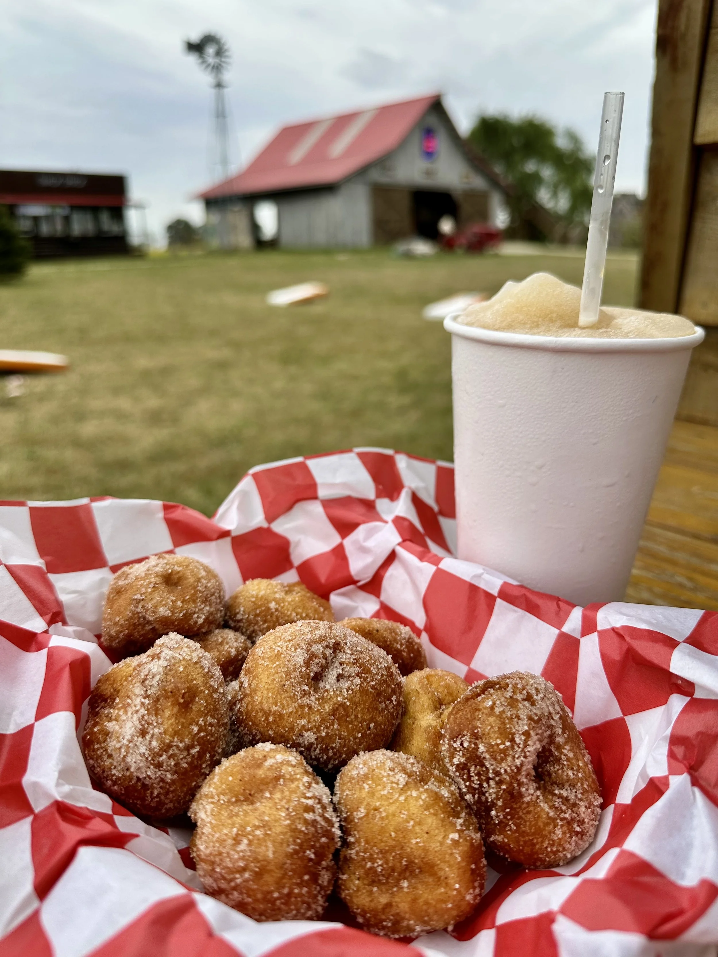 Basket of fried food balls on red and white checkered paper with a foam cup of frozen drink with a straw in front of a barn and windmill outdoors.