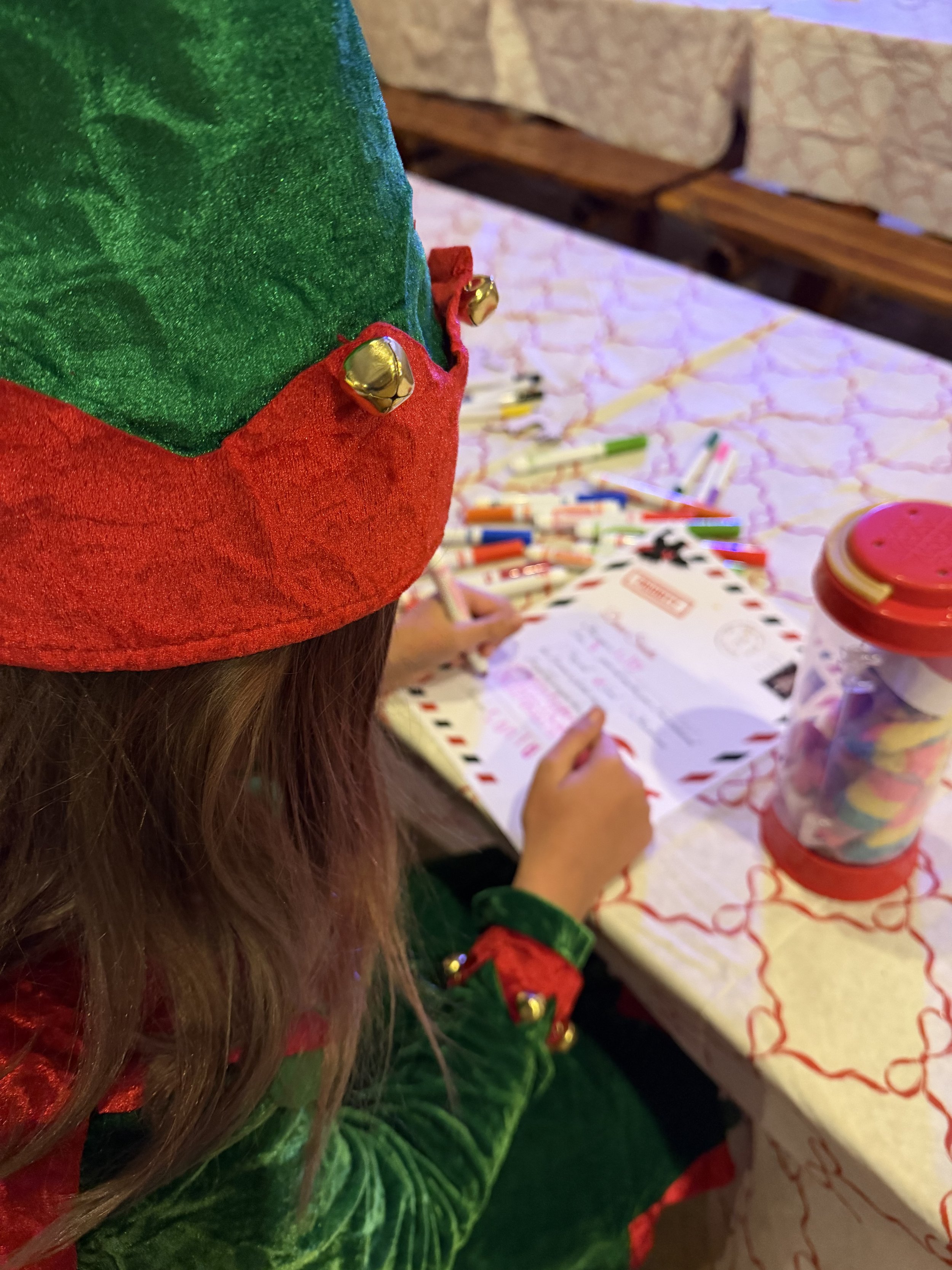 Child dressed in Christmas elf costume sitting at a table decorated with Christmas-themed tablecloth and surrounded by holiday decorations and treats.