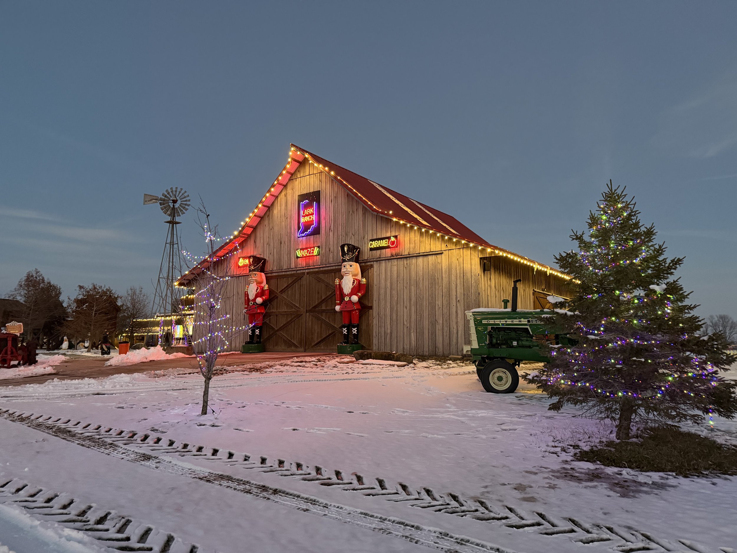 A wooden barn decorated with Christmas lights, Nutcracker figures, and neon signs reading "Lark Ranch," "Maze," and "Caramel". There is a Christmas tree with lights on the right side and snowy ground with tire tracks in the foreground. A windmill and some trees are visible in the background during dusk or evening.
