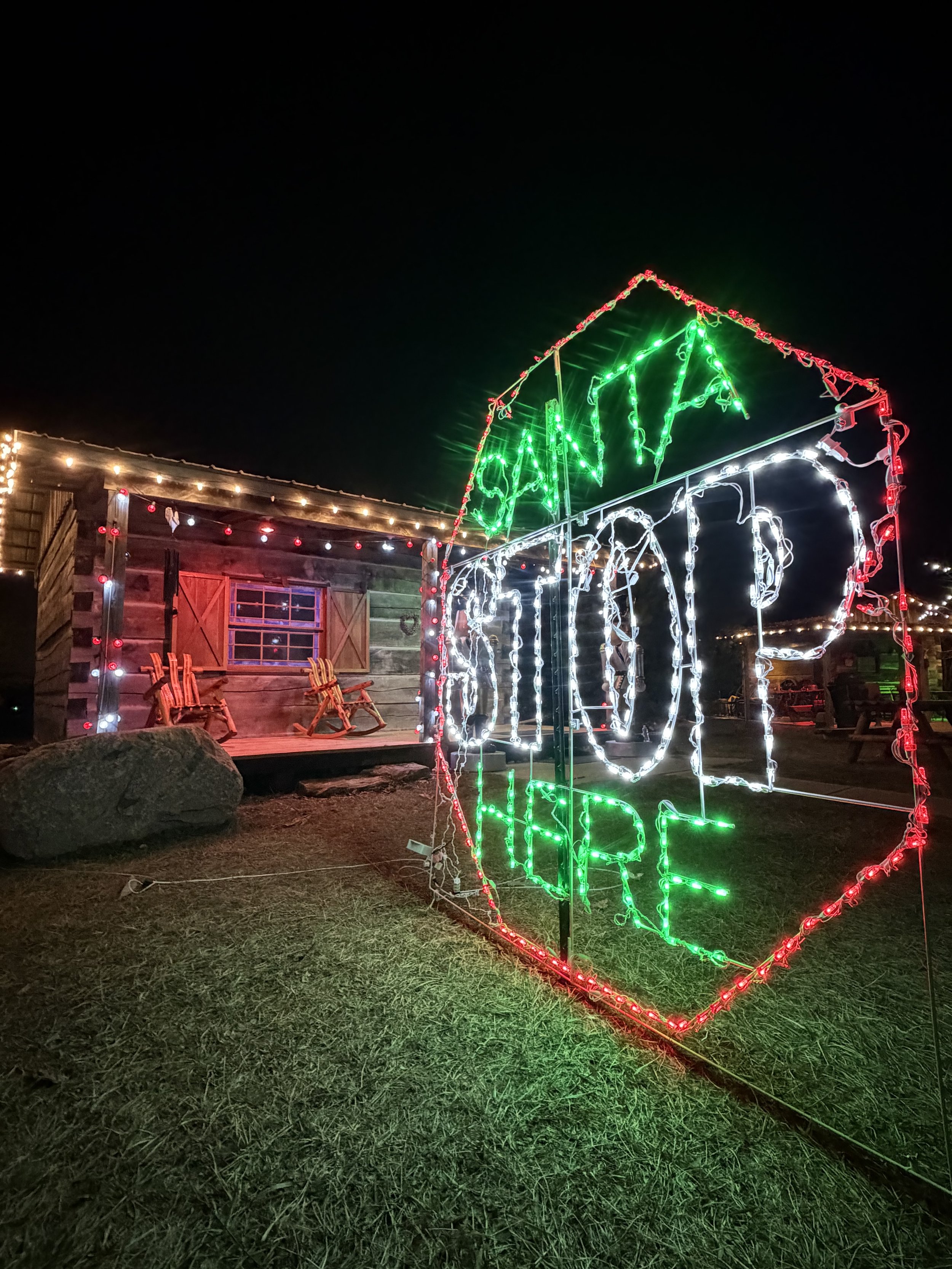 Nighttime scene with holiday lights, including a large white lit Christmas tree on the right and a rustic wooden building with string lights and chairs on the left.