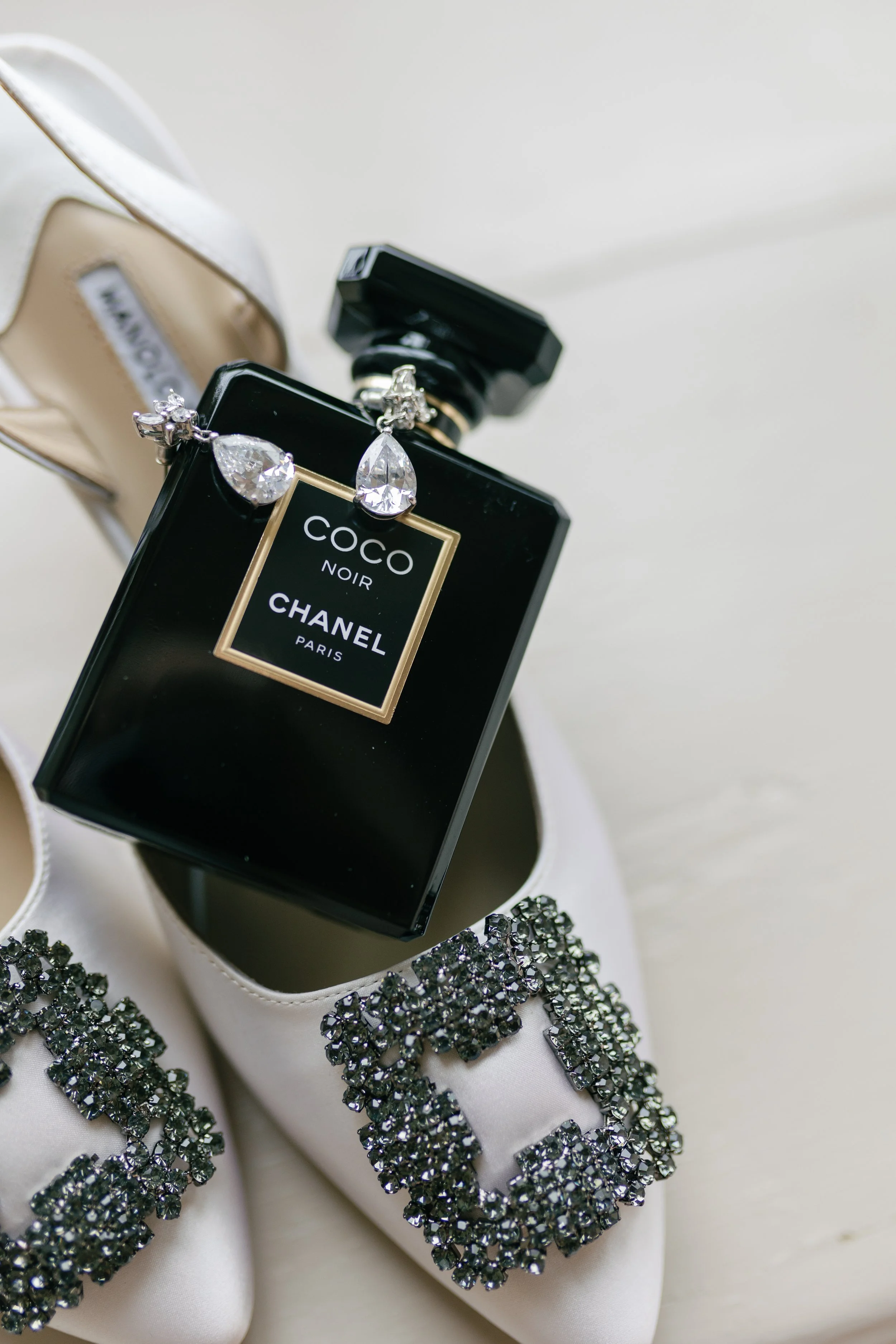 White satin wedding shoes with black rhinestone embellishments, a bottle of Chanel Coco Noir perfume, and a pair of diamond earrings.