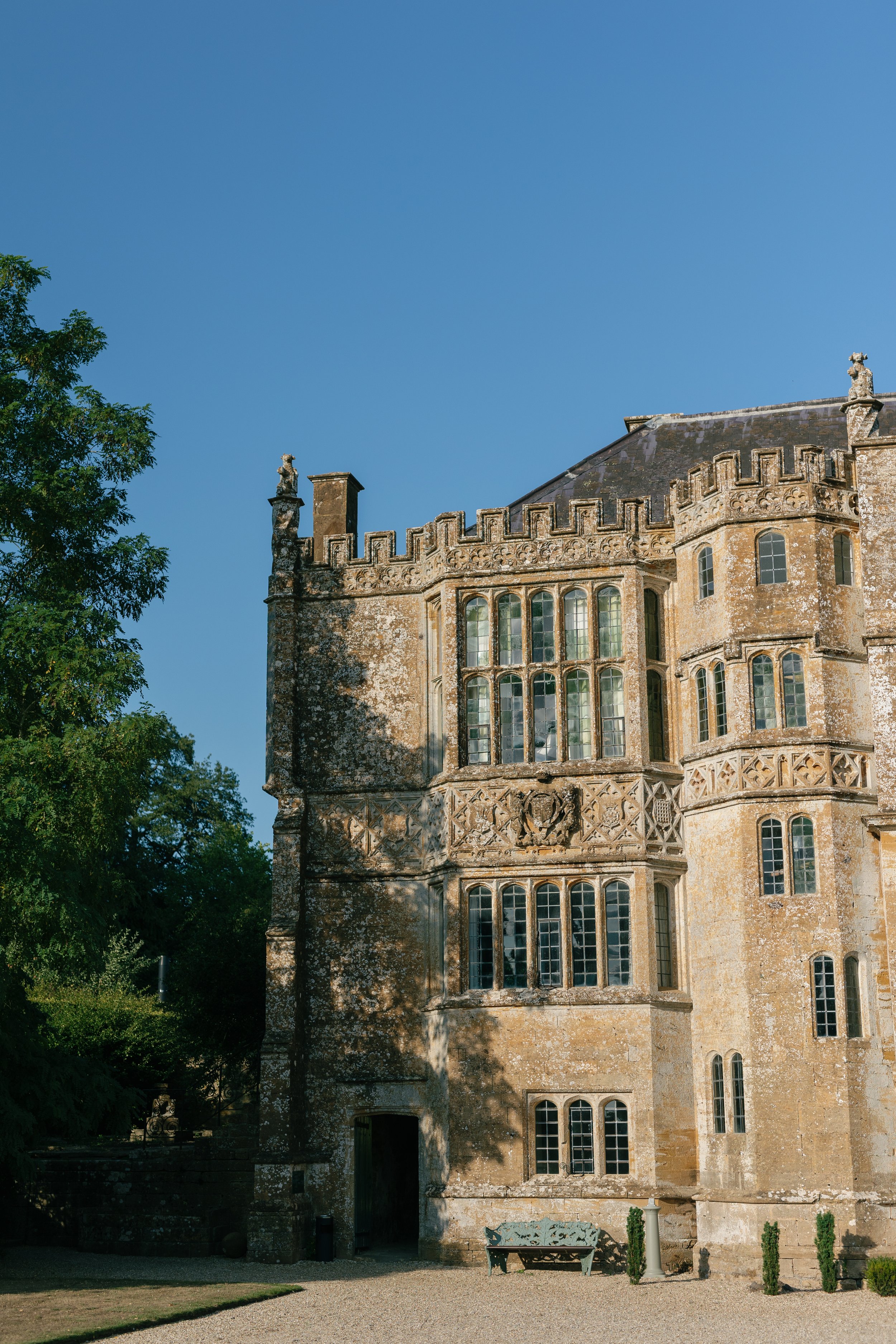 A historic stone castle with detailed architecture and large windows, surrounded by greenery and a clear blue sky.