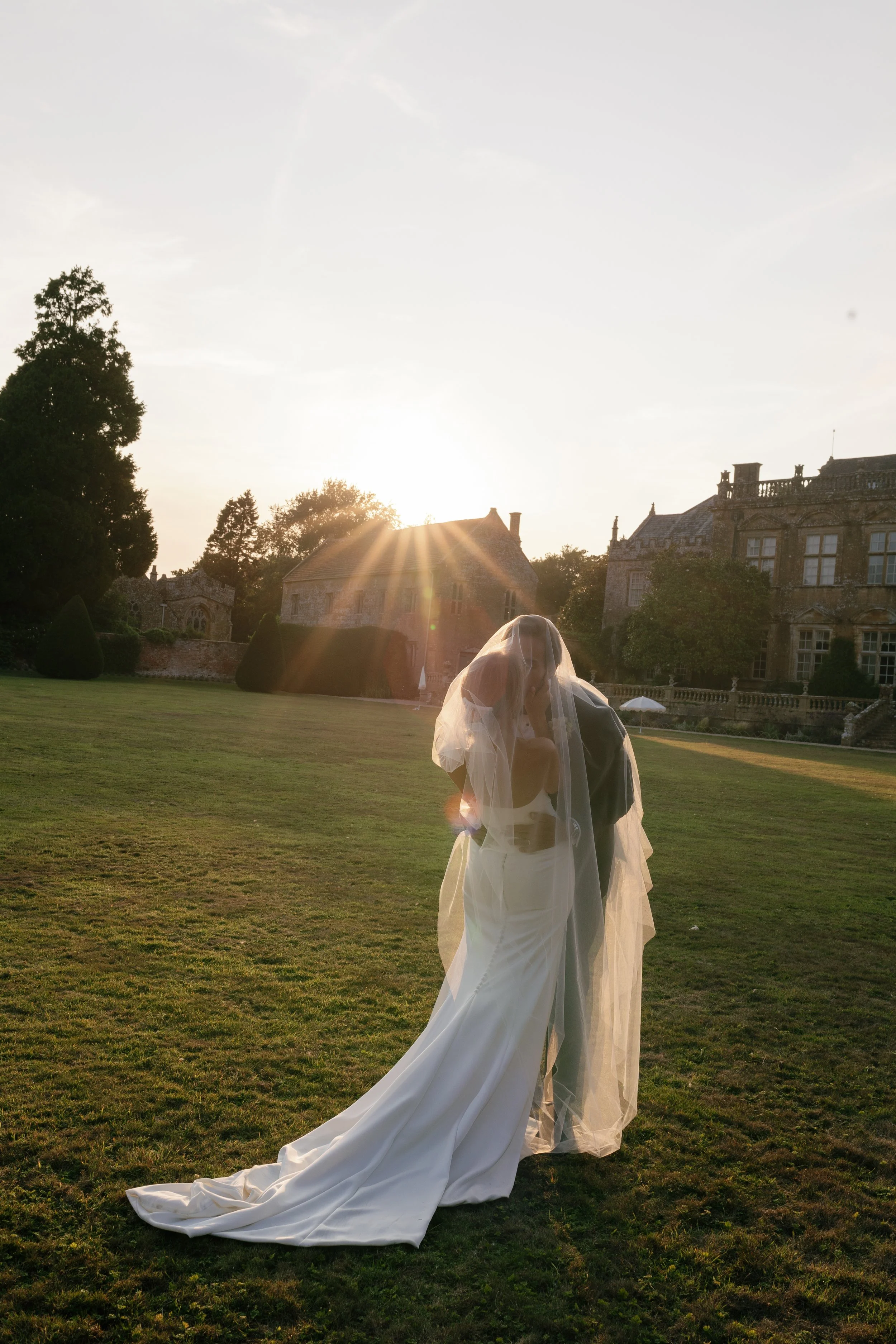 A bride and groom share an intimate moment outdoors during sunset on a grassy lawn, with a large historic building in the background.