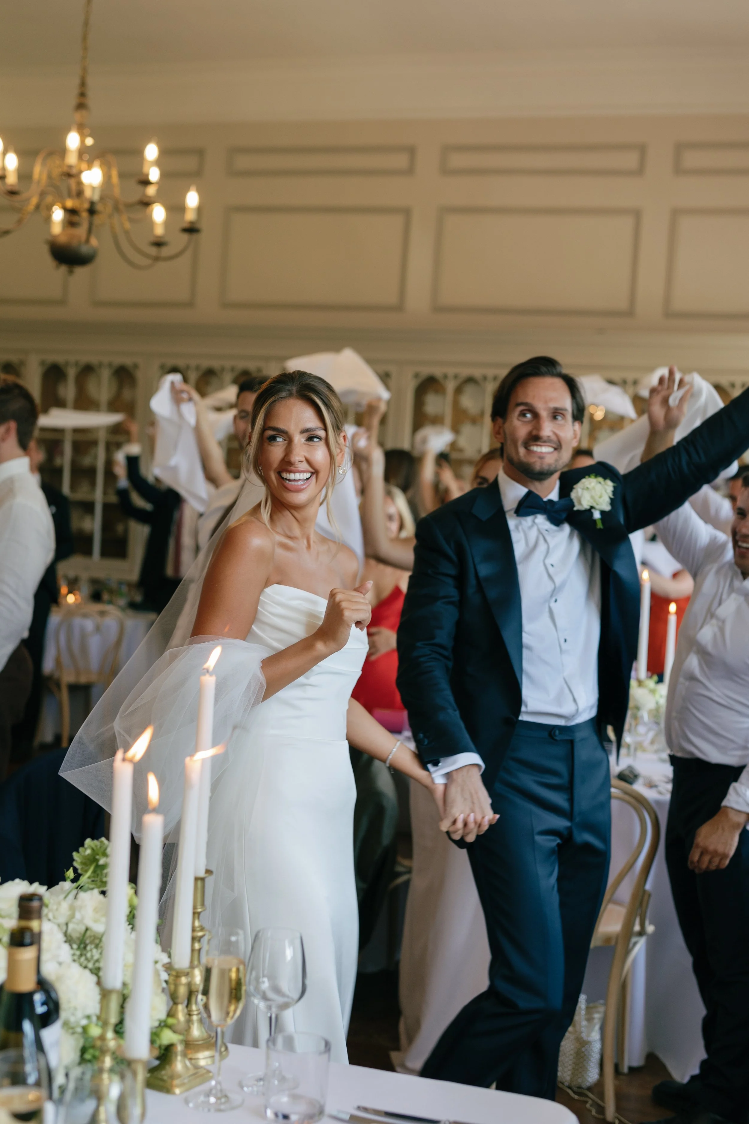 Bride and groom holding hands and smiling during their wedding reception surrounded by guests, tables, and candles.