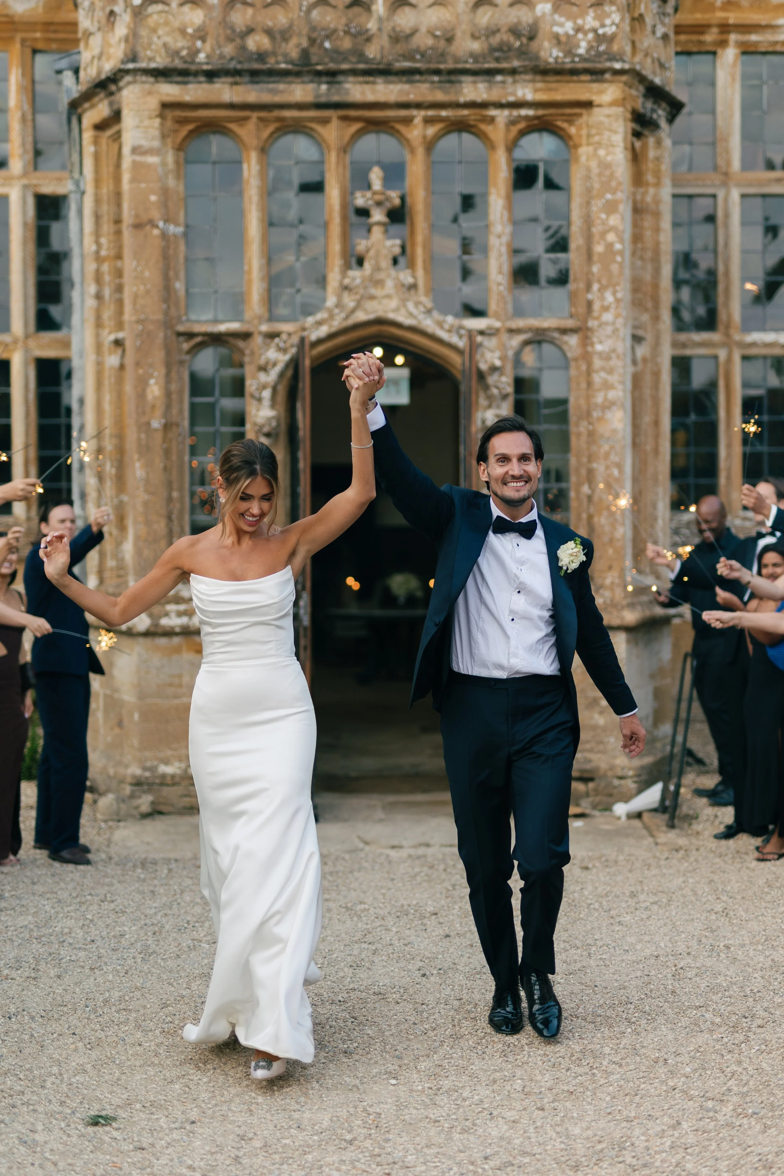 A newlywed couple walking hand in hand, smiling and celebrating outside a historic building with wedding guests surrounding them.