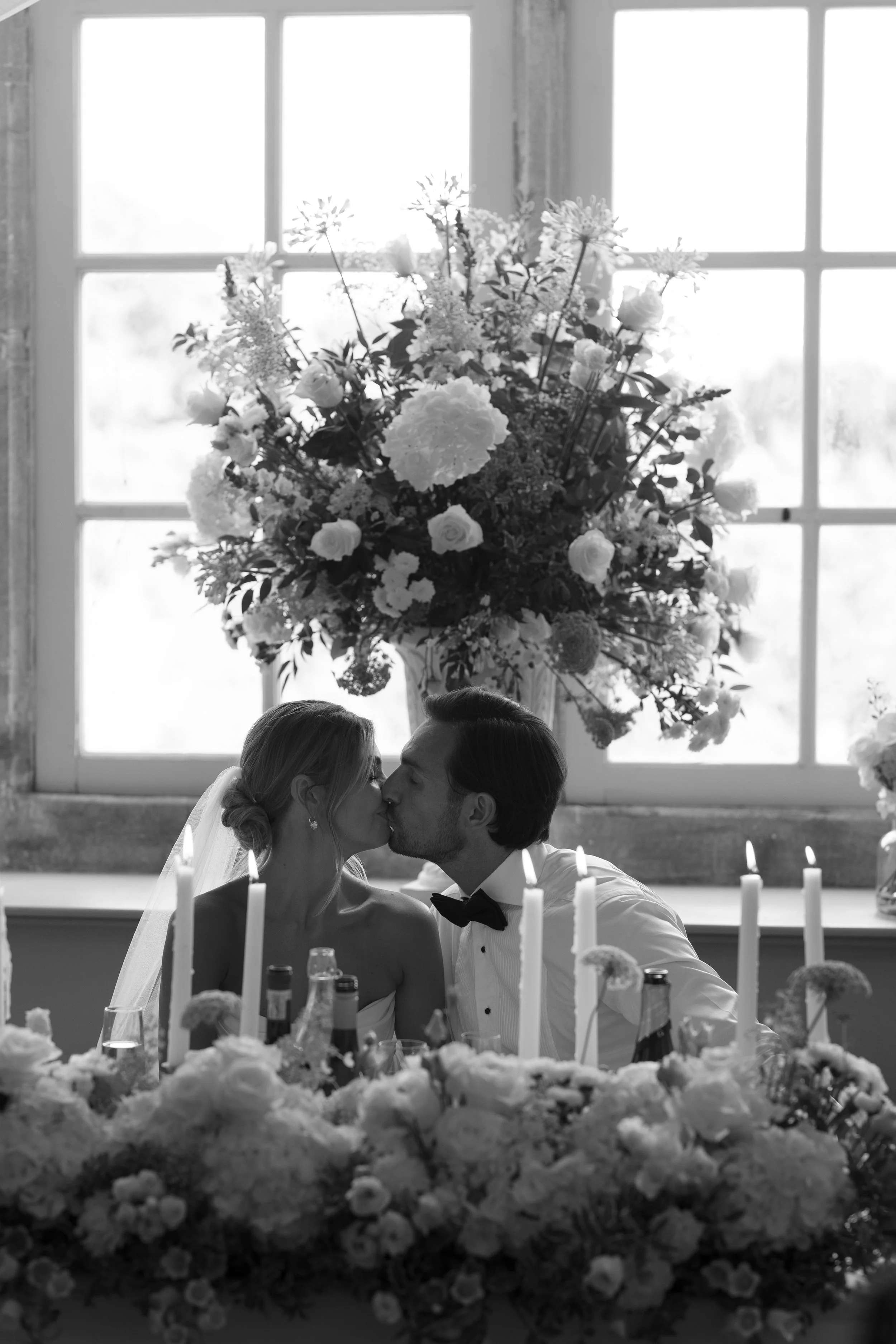 A black and white photo of a bride and groom kissing at their wedding reception, seated at a table decorated with flowers and candles, with large floral arrangements in the background in front of a window.