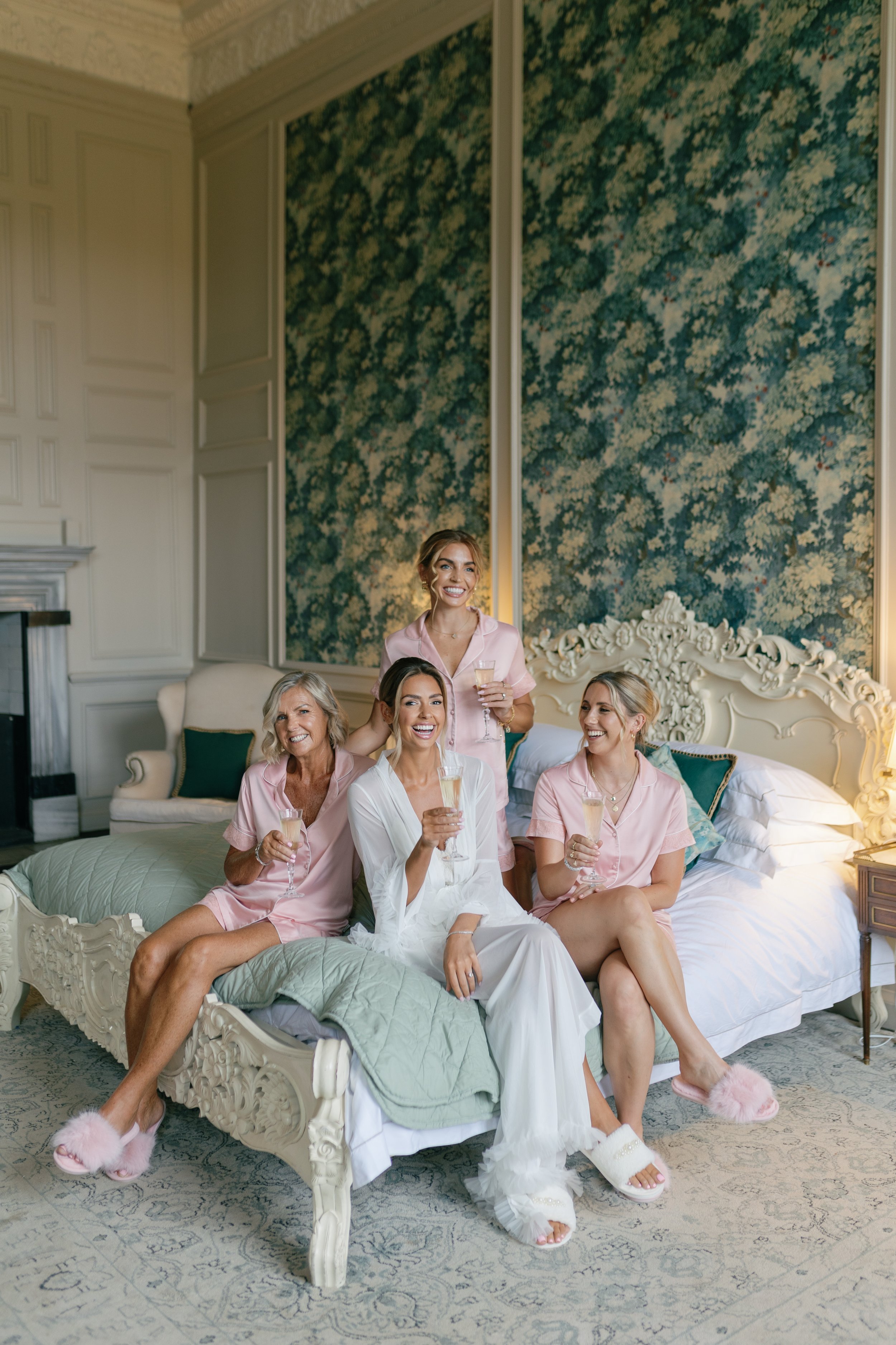 Five women in pajamas sitting and standing on a bed enjoying champagne in a luxurious bedroom.