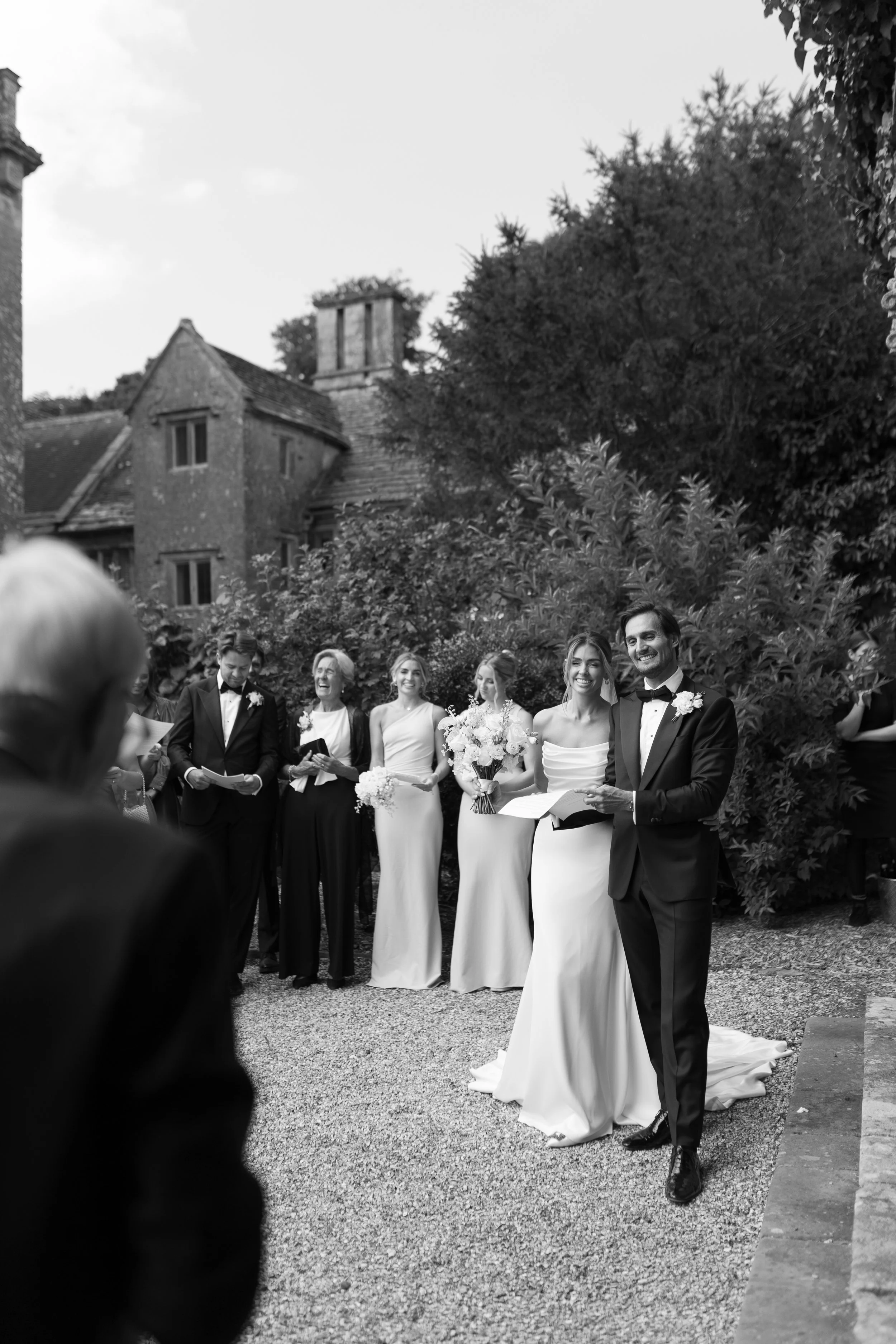 A wedding ceremony outdoors with a group of smiling people, including the bride and groom, standing on gravel in front of a large bush and an old brick building with a chimney in the background. The groom wears a tuxedo, and the bride is in a white w
