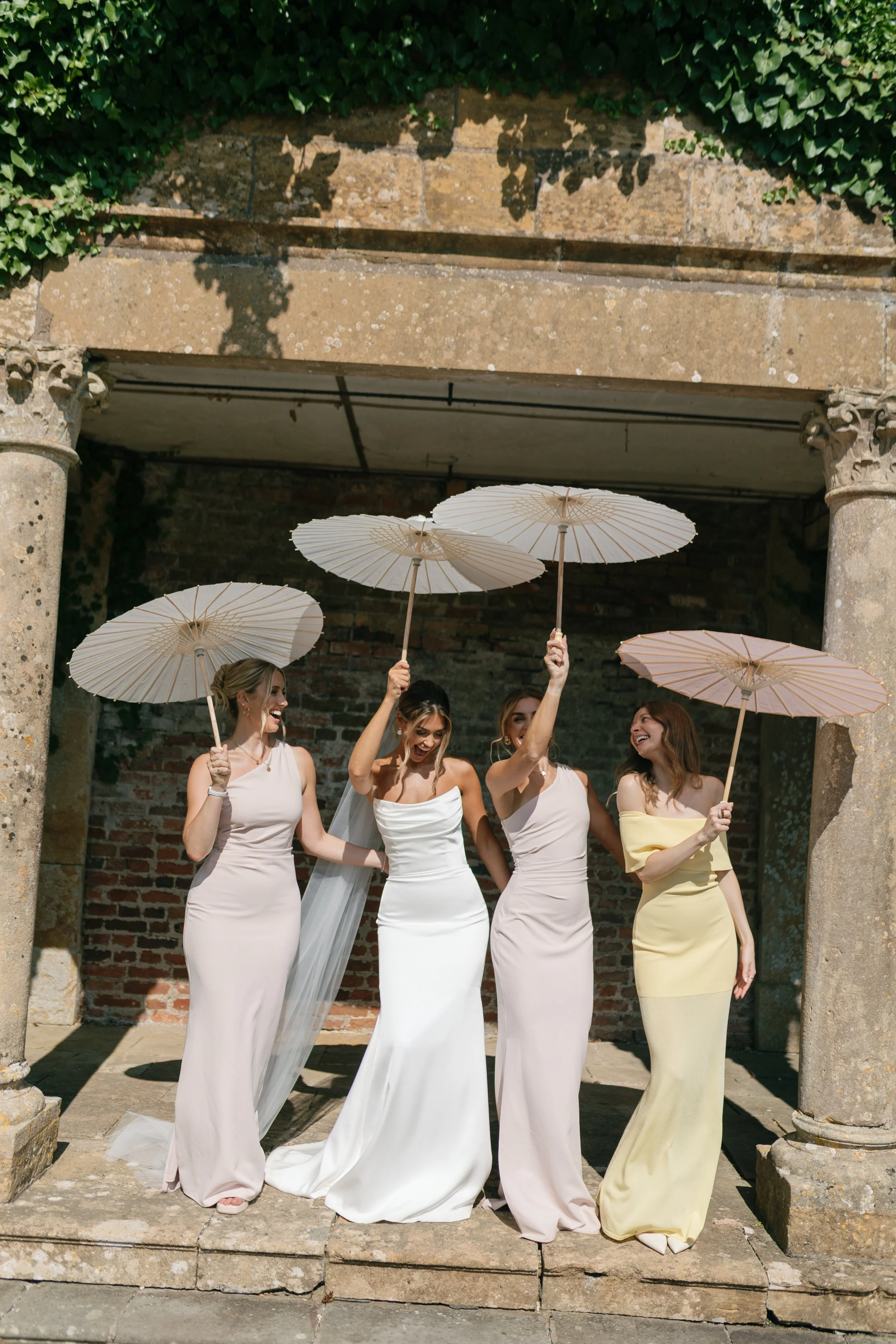 Four women in elegant dresses holding parasols, celebrating outdoors under a Covered archway.