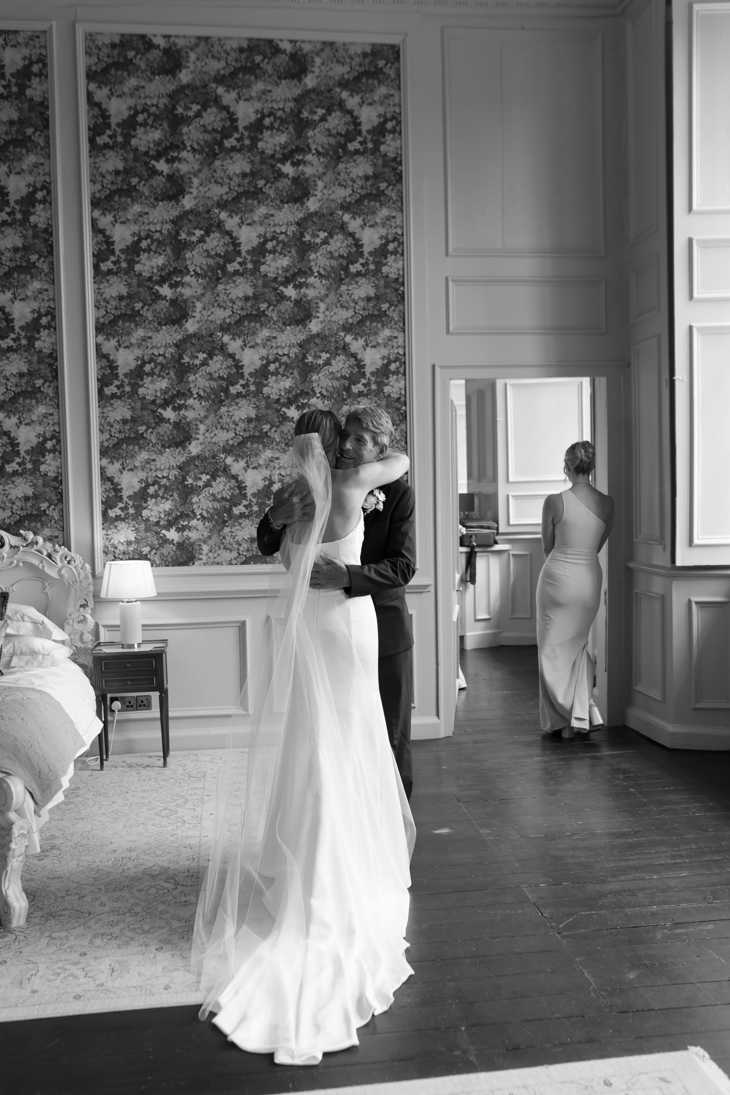 A bride and groom hugging in a vintage-style room, with a woman in a one-shoulder gown in the background, all in black and white.
