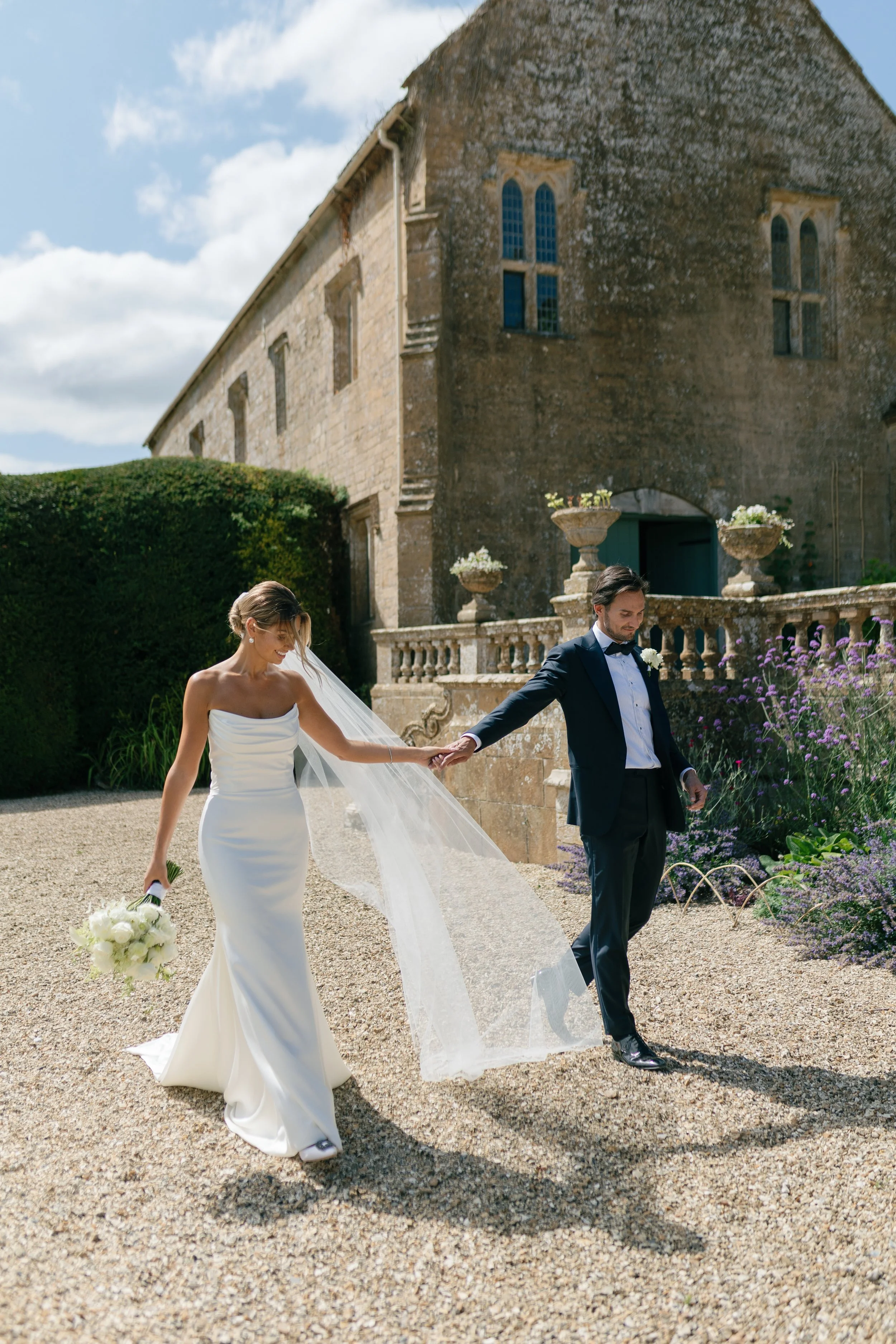 Bride and groom holding hands outside a stone building with flowering plants and a gravel path
