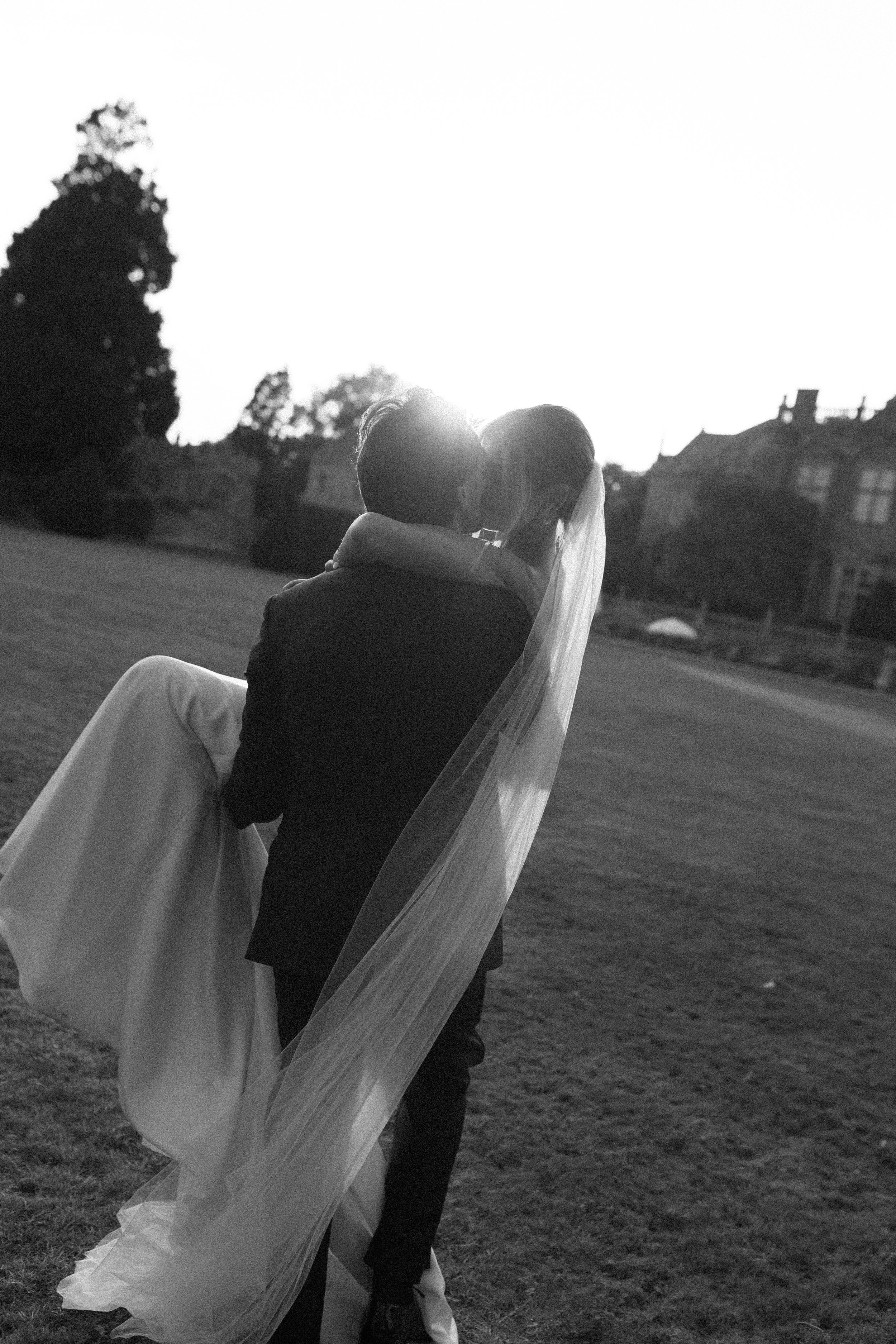 A black-and-white photo of a bride and groom embraces outdoors at sunset, with the bride's veil flowing.