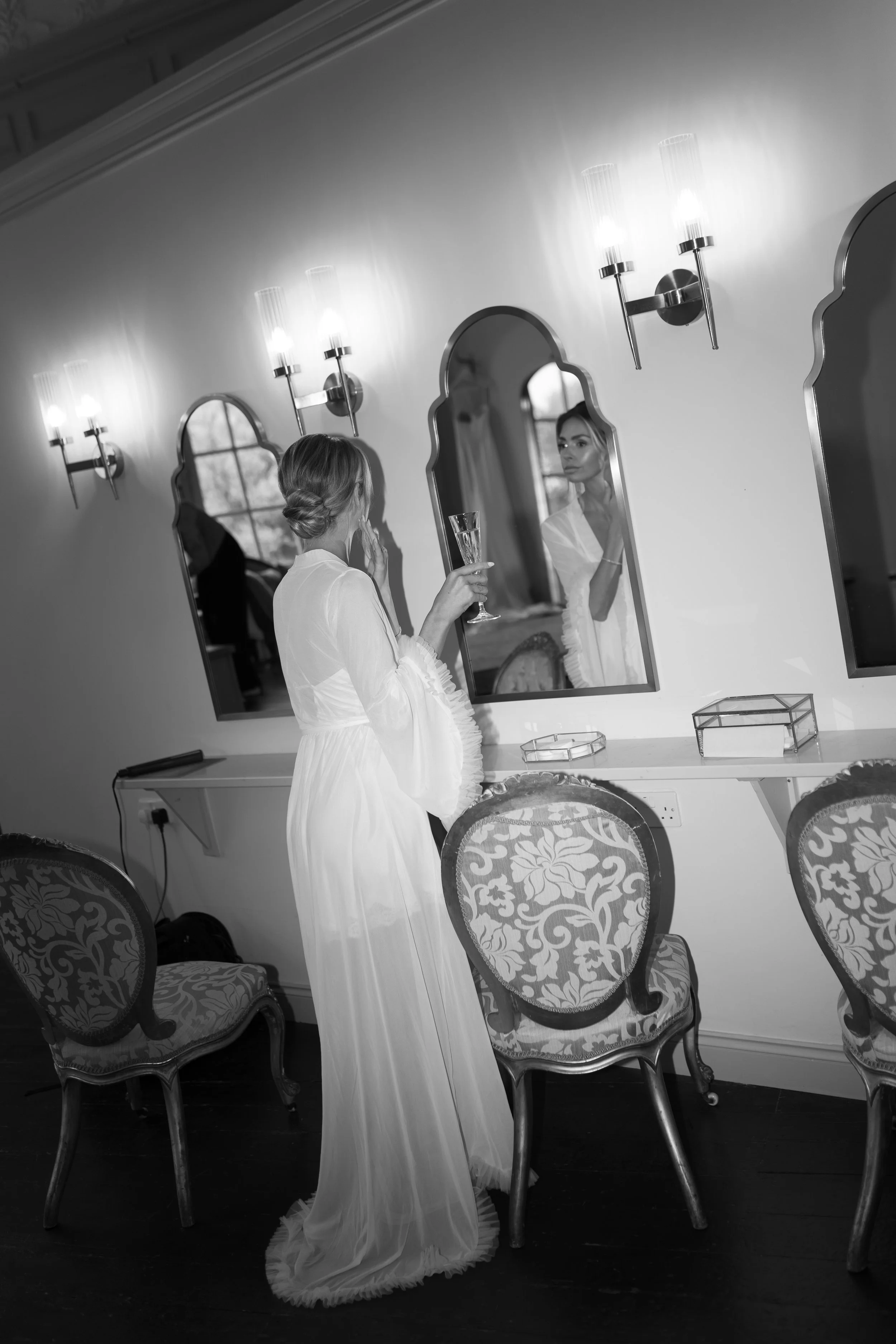 A woman in a long, white dress stands in front of a mirror holding a glass of champagne, looking at her reflection.