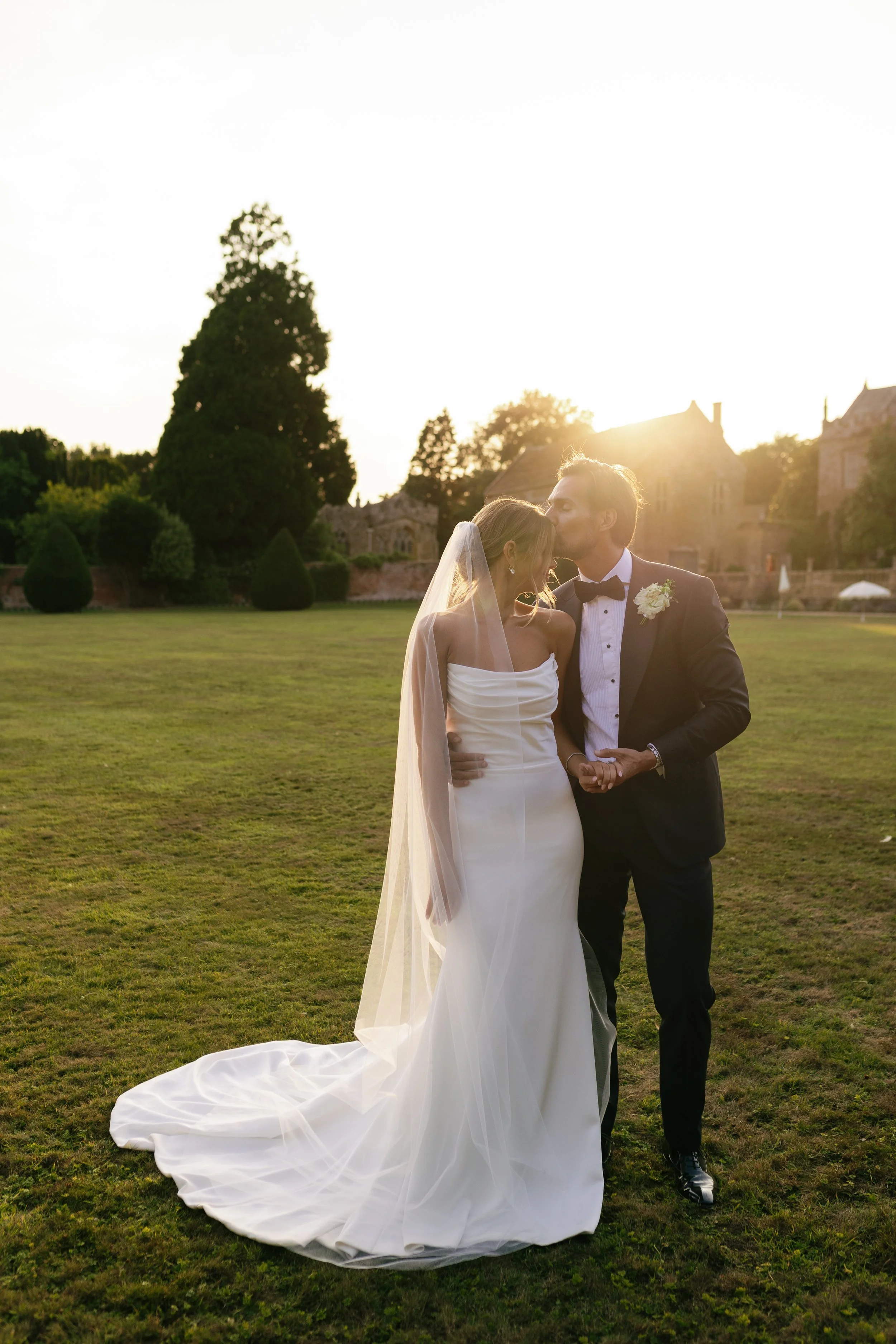 A bride and groom sharing a kiss during their wedding, standing outdoors on a grassy area with a historic-looking building in the background at sunset.