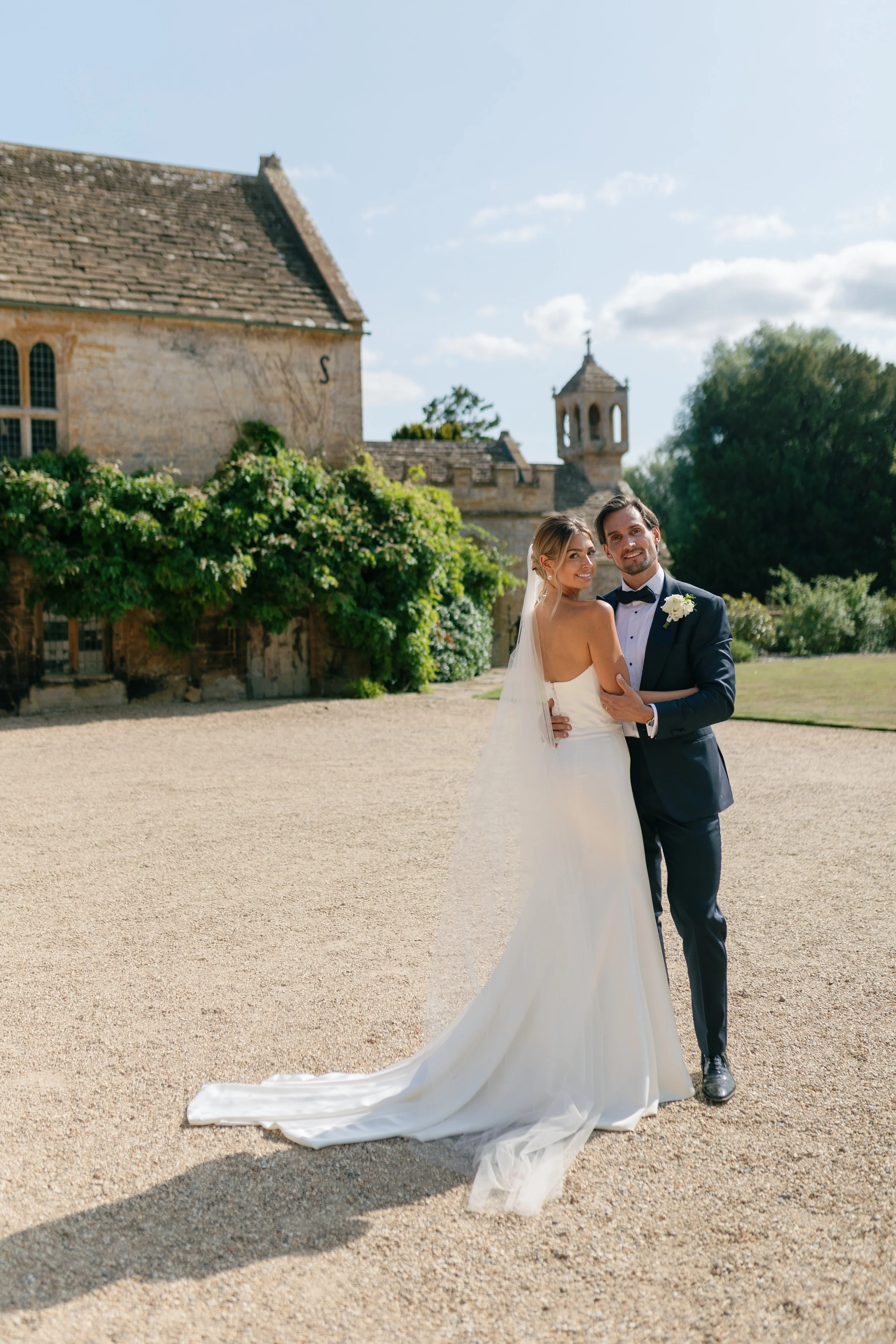 A bride and groom standing outdoors on a sunny day, smiling and embracing in front of a historic stone building and lush greenery.