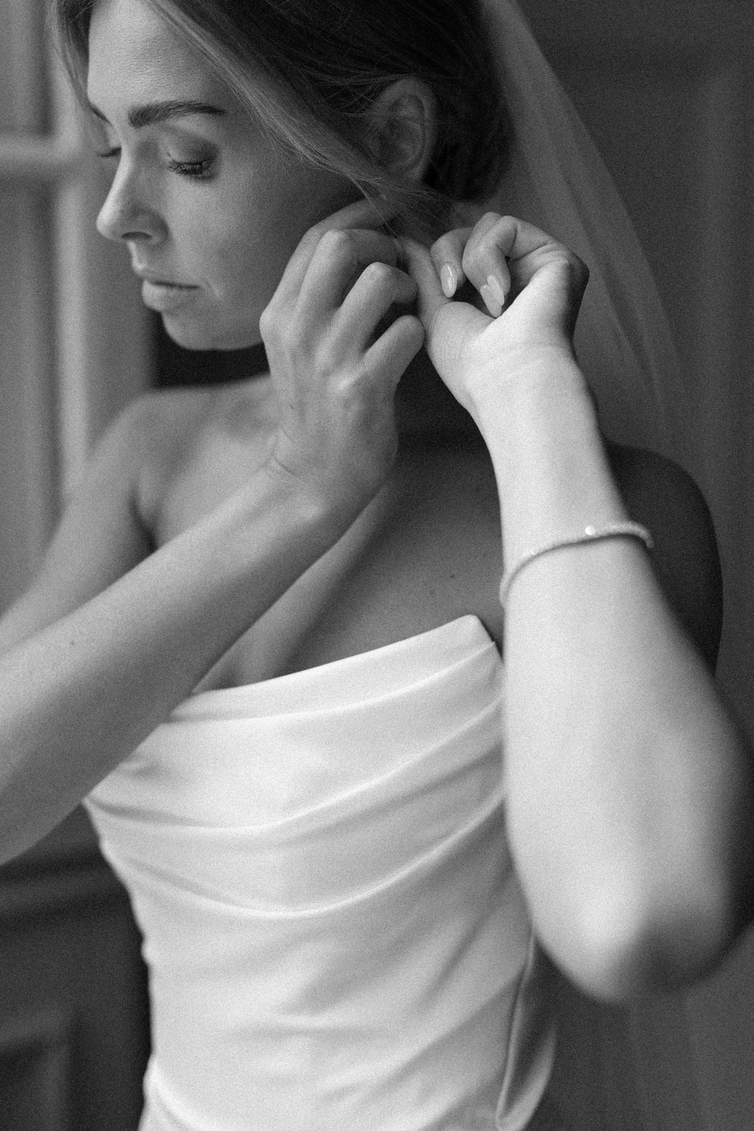 A woman adjusting her earring, dressed in a strapless satin wedding gown, with a veil and a bracelet on her wrist.