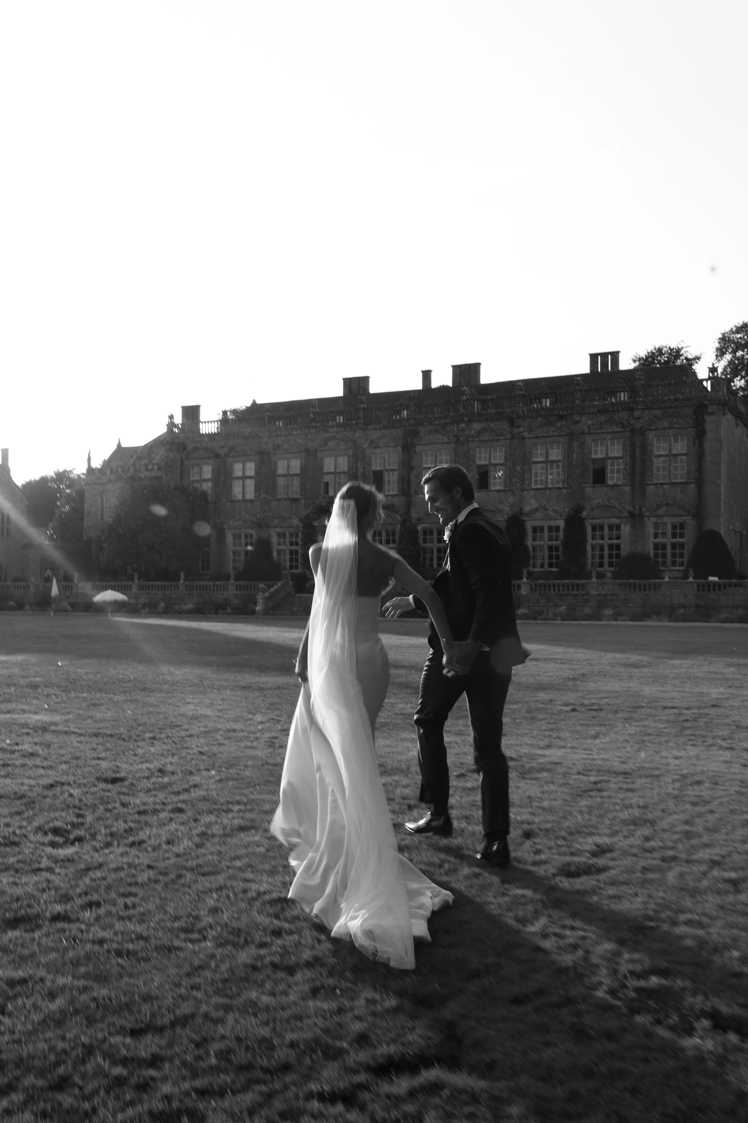Black and white photo of a newlywed couple dancing outdoors on a grass lawn, with a large historic building in the background during sunset.