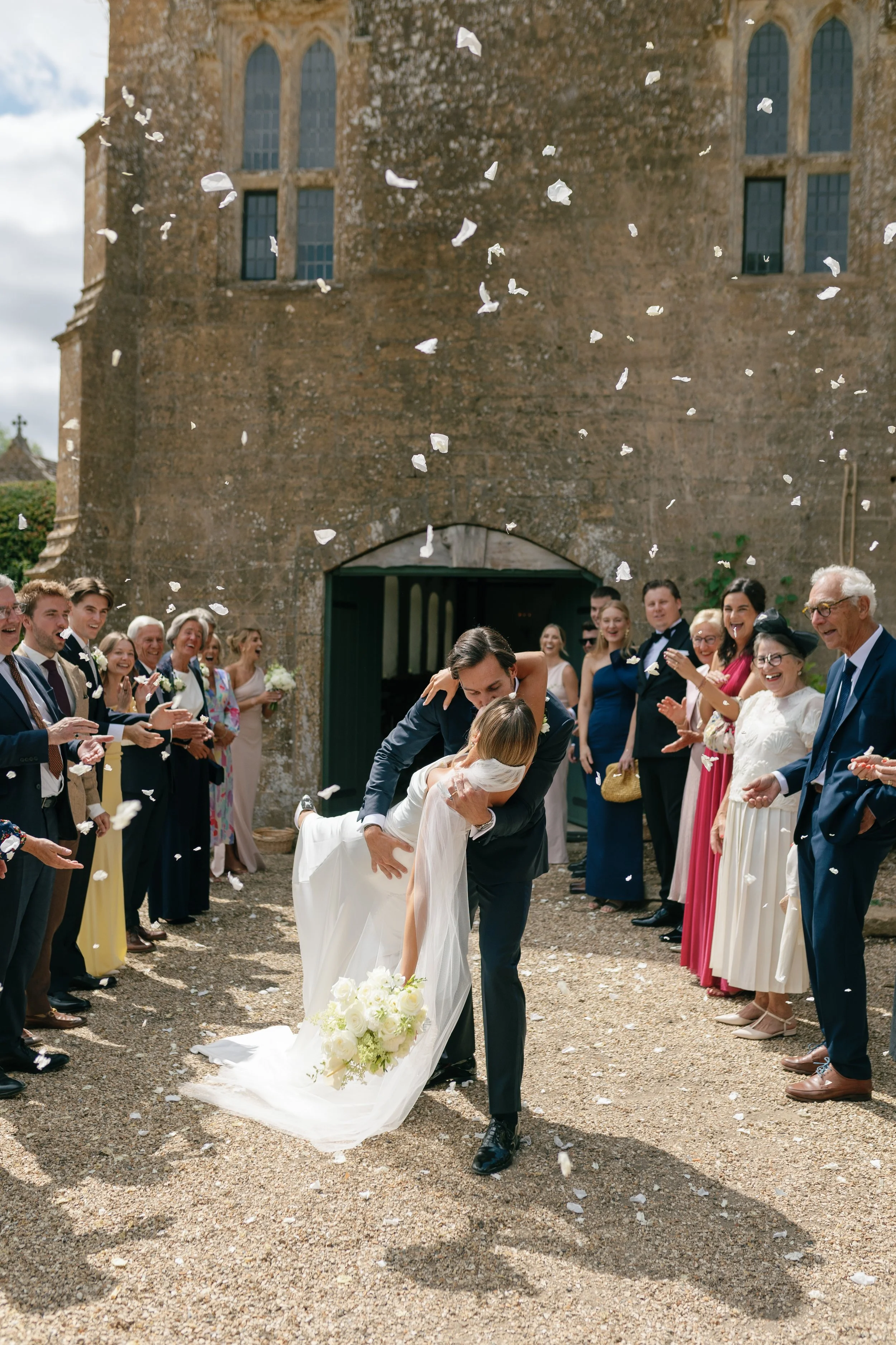 A bride and groom sharing a dance outside a church with guests celebrating around them, throwing white flower petals in the air.