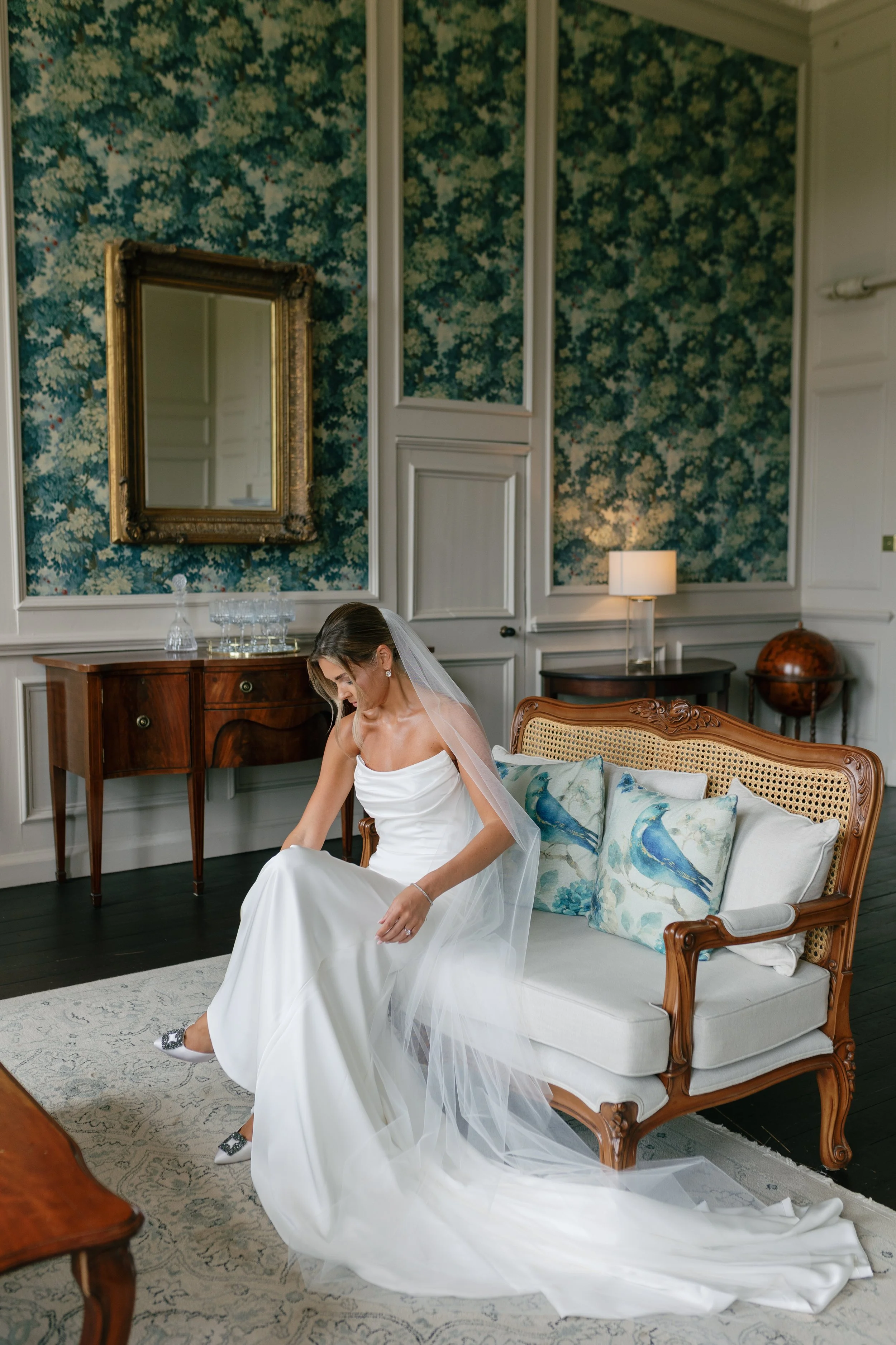 Bride in a white wedding dress and veil sitting on a wooden vintage sofa with blue bird pillows in an elegant room with floral wallpaper, a mirror, a sideboard with glassware, and a table lamp.