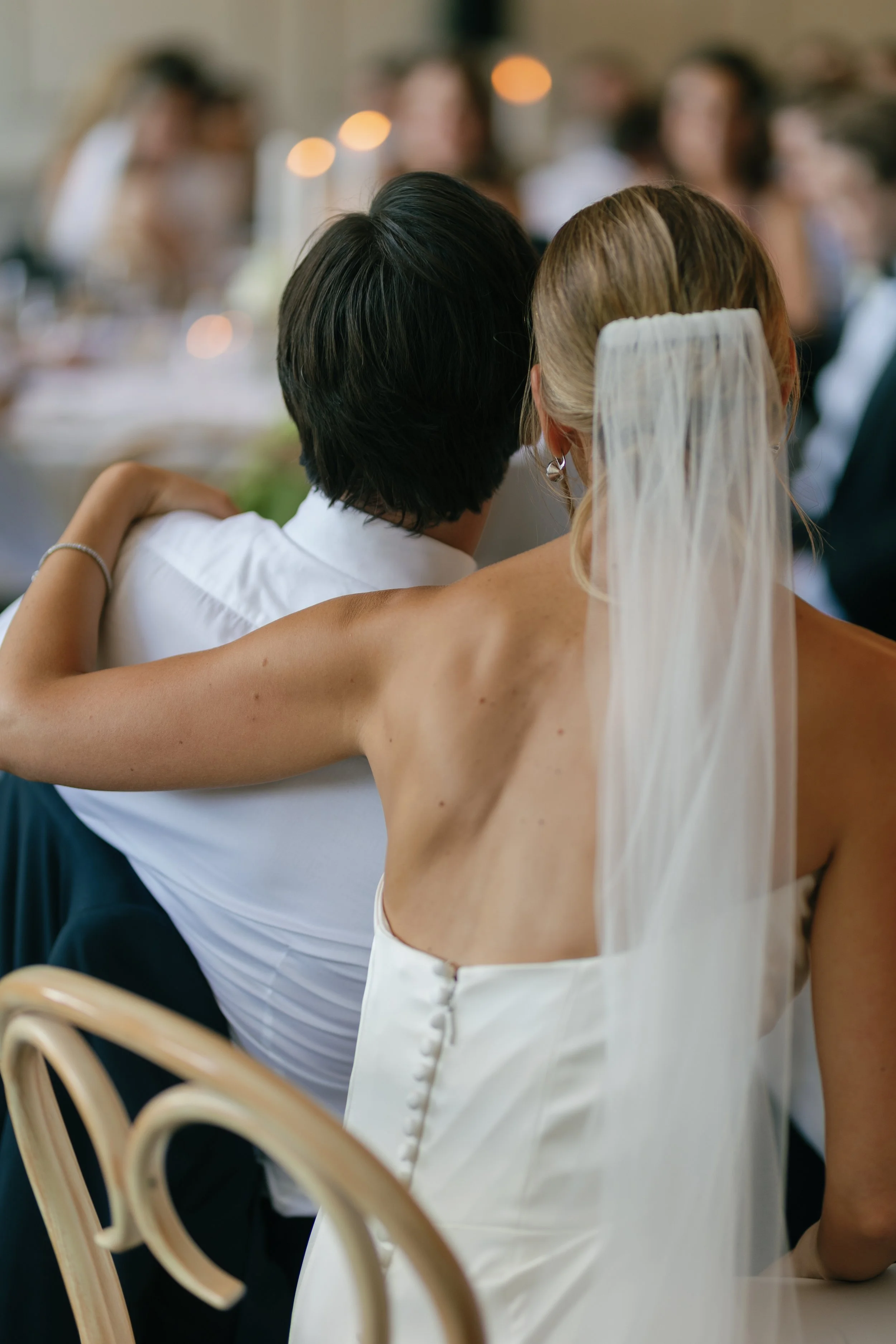 A bride with a veil and a white strapless wedding dress sitting next to a groom in a white shirt at a wedding reception.