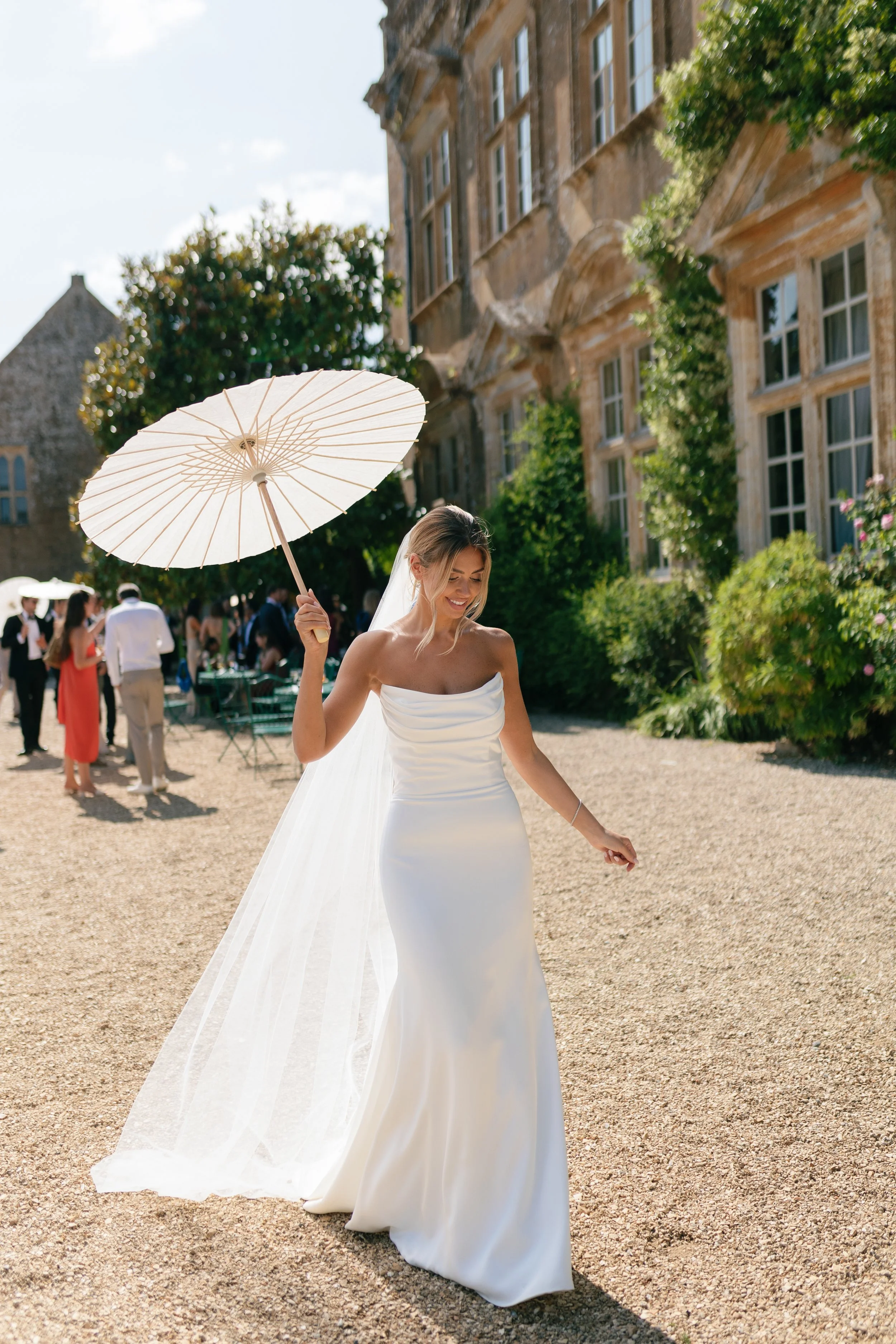 A bride in a white satin wedding dress holding a white parasol while walking outdoors on a gravel path, with a historic building and some guests in the background.