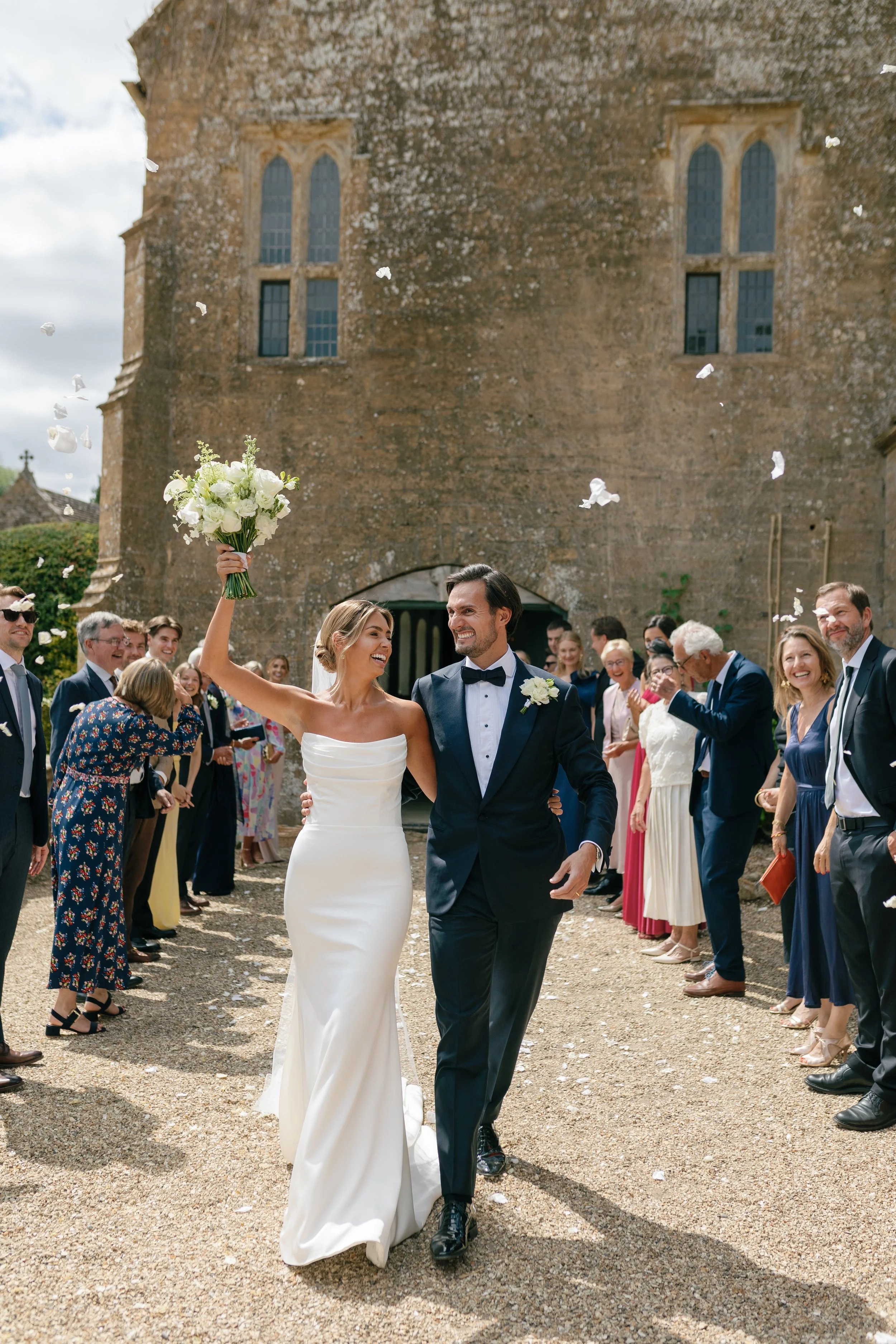 Bride and groom walking outside a church with guests throwing flower petals in the air, celebrating their wedding.