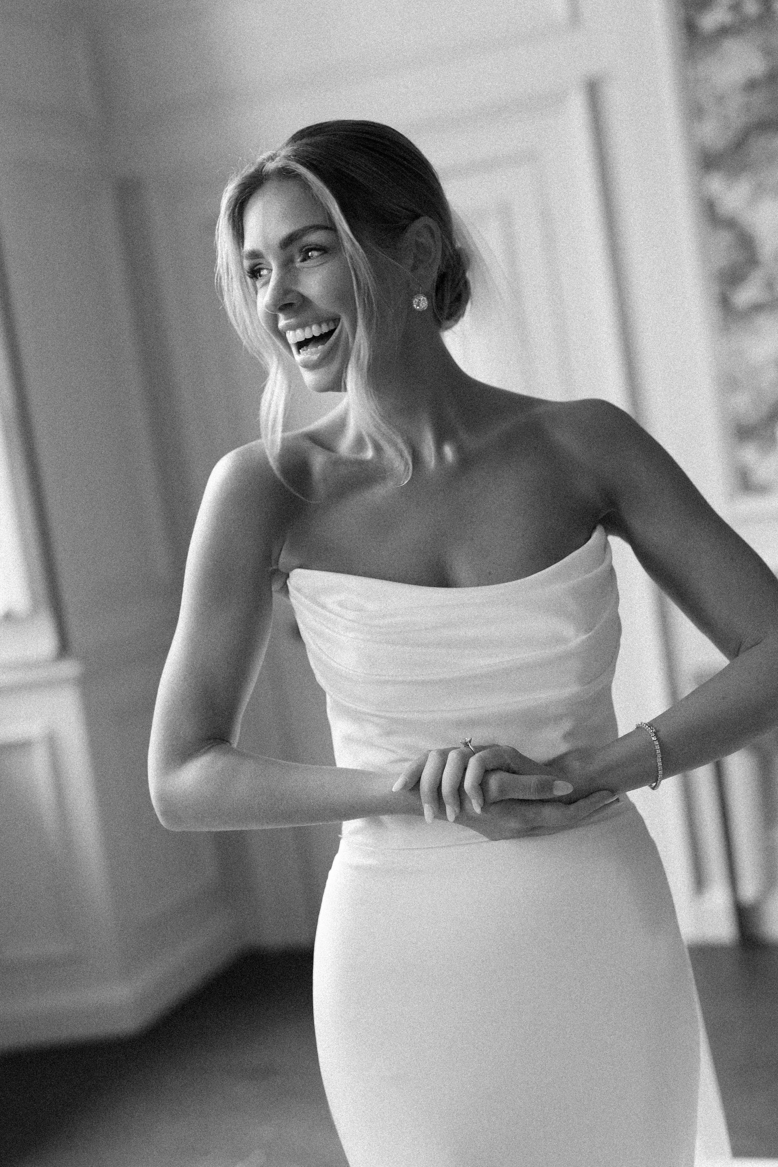 A smiling woman in a strapless dress with jewelry, standing indoors, captured in black and white.