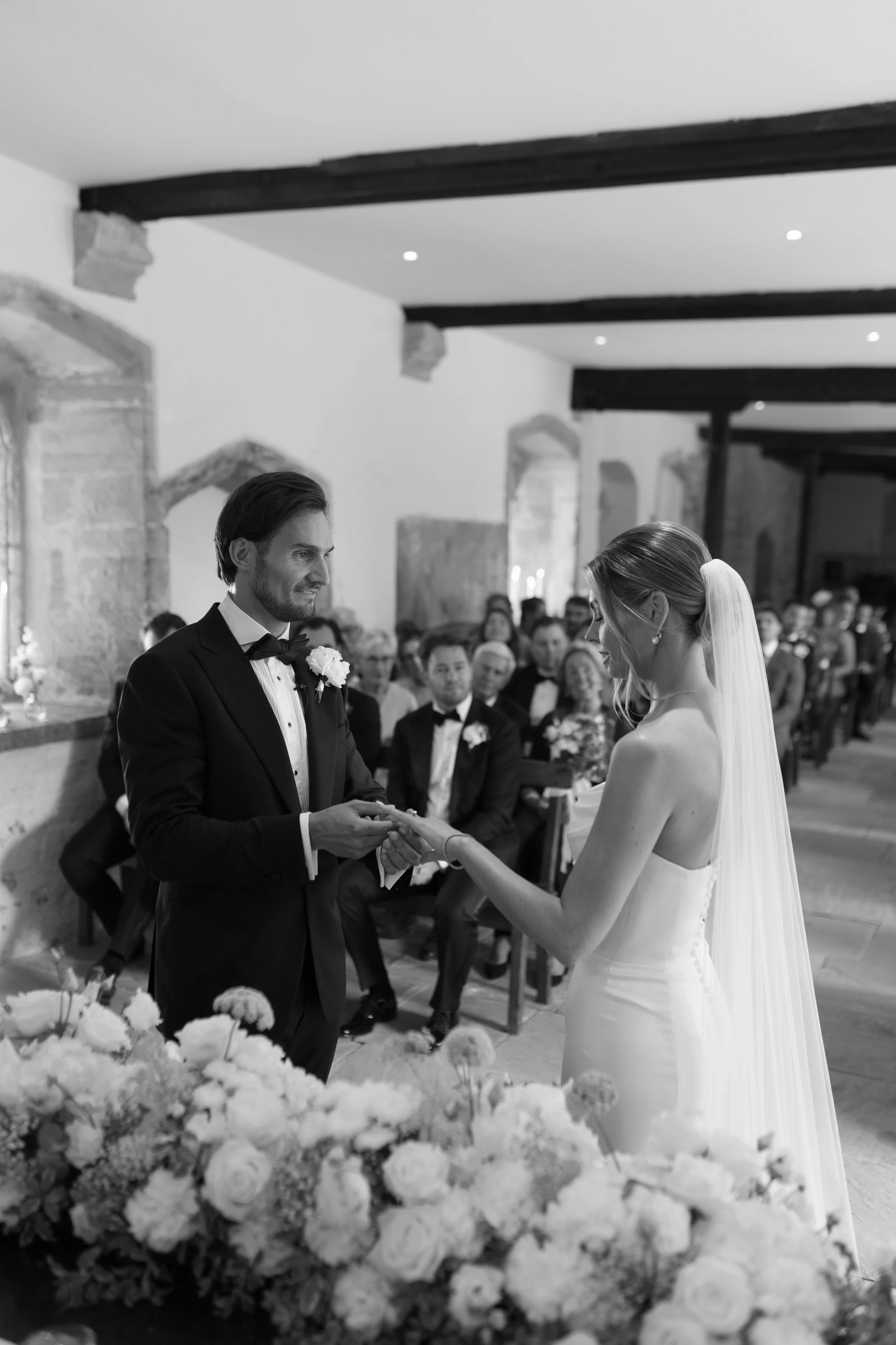 A black and white photo of a wedding ceremony with a groom exchanging rings with a bride. The groom is wearing a tuxedo and the bride is in a strapless wedding gown with a veil. Guests are seated in the background.