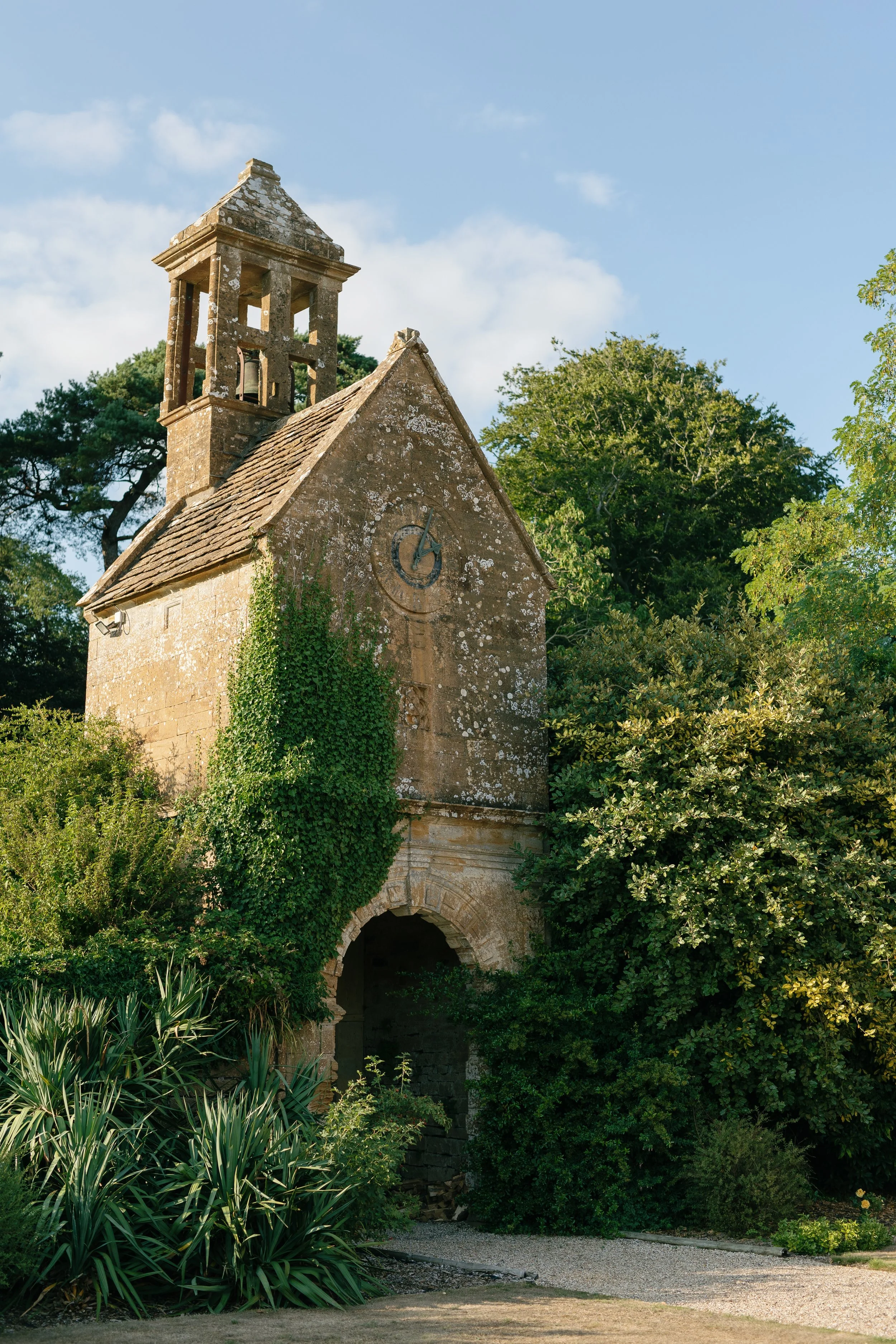 An old stone clock tower with ivy growing on it, surrounded by lush green bushes and trees, under a blue sky with some clouds.