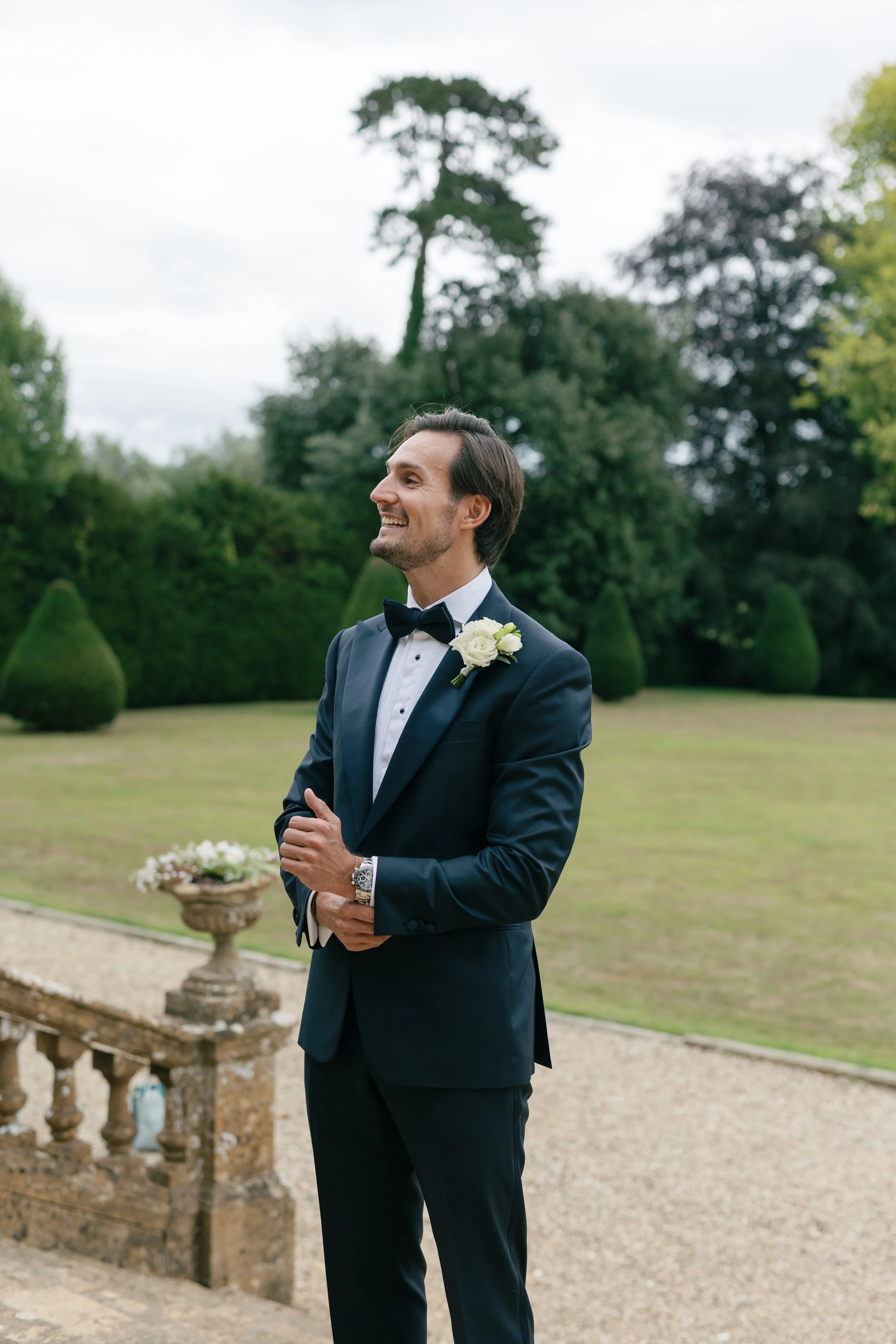 A man in a tuxedo with a white flower boutonniere, smiling outdoors in a garden setting.