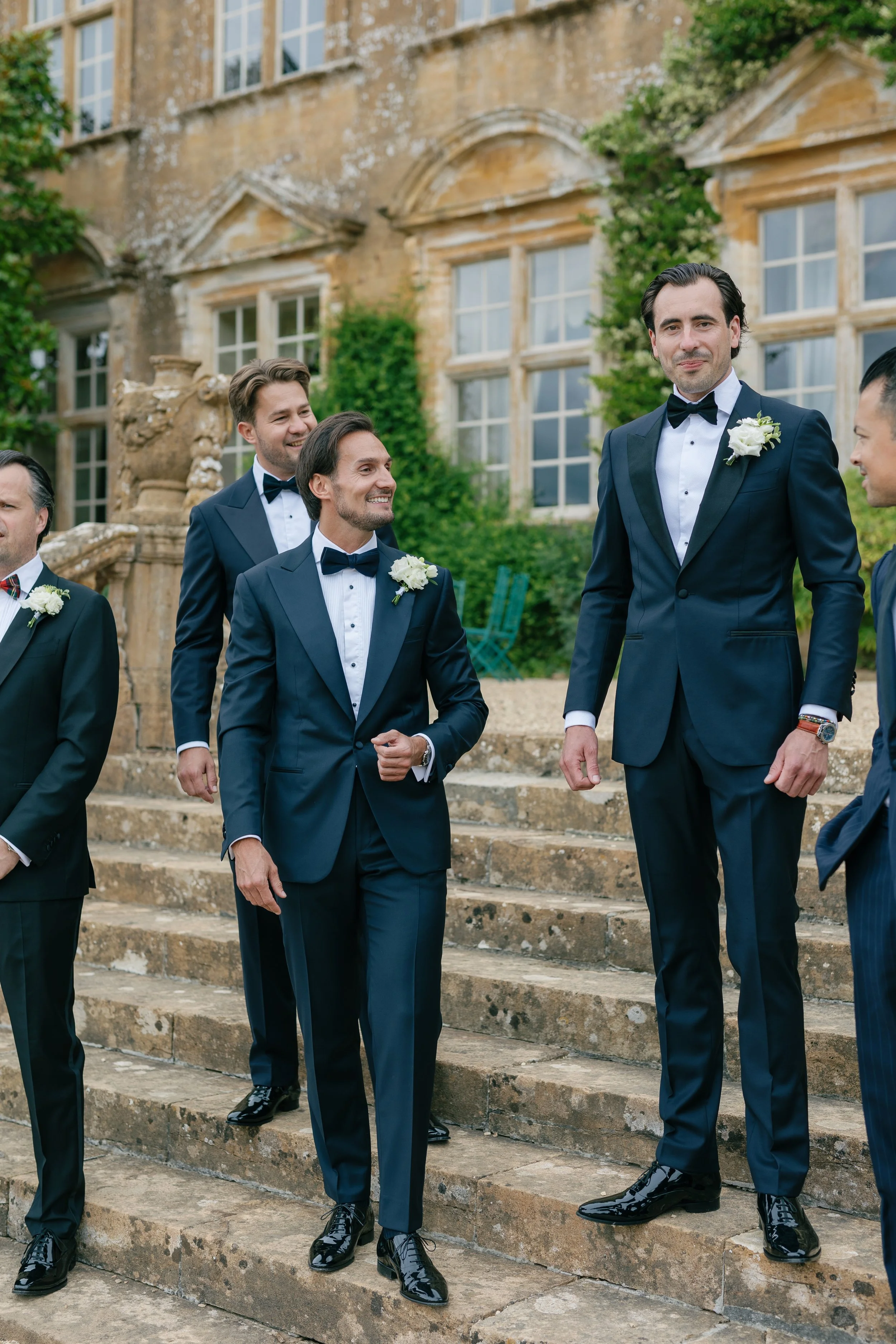 Group of men dressed in formal navy tuxedos with bow ties, standing on stone steps outside an old building with large windows and greenery.