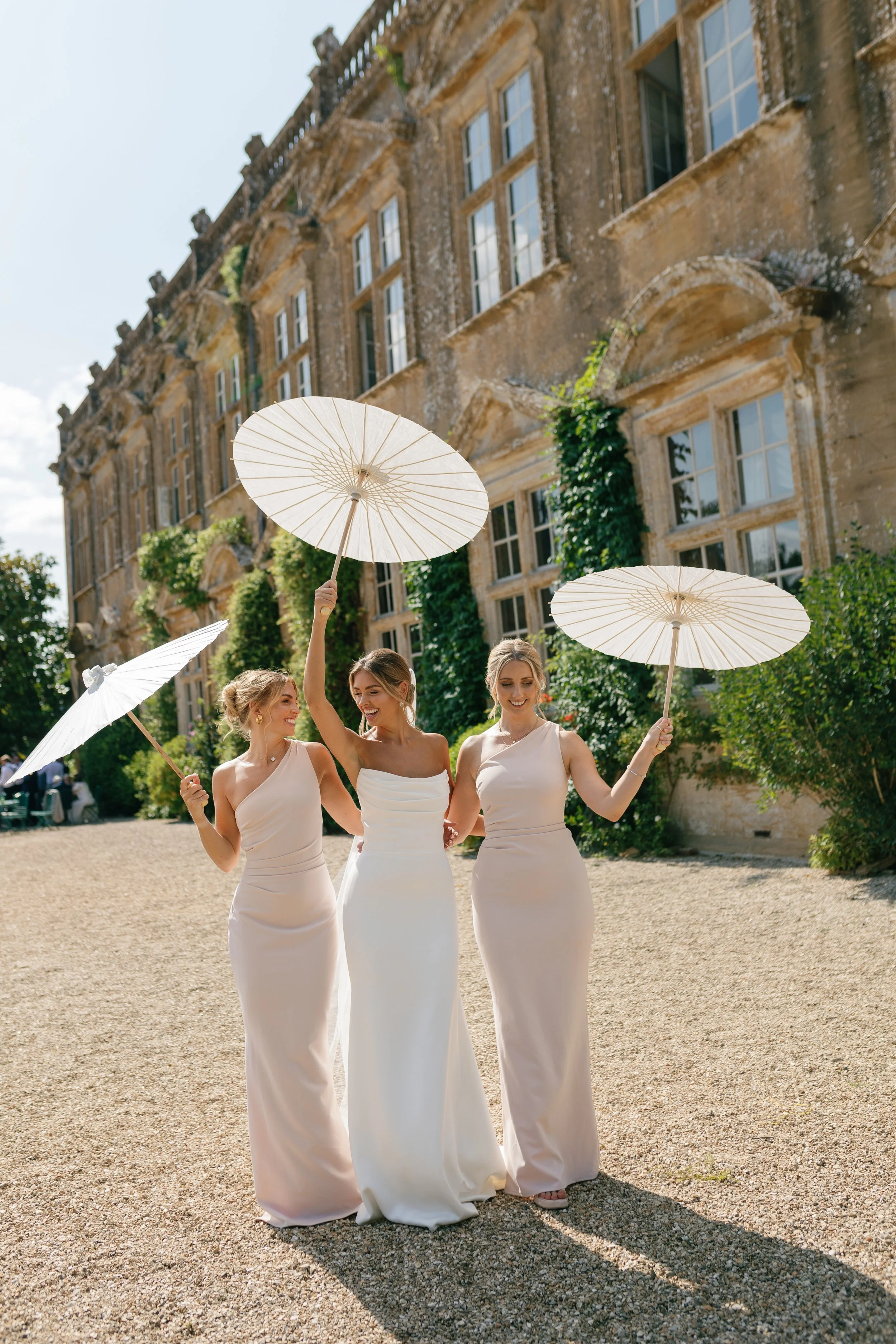 Three women in white dresses holding parasols, smiling and posing outdoors in front of an old stone building covered with greenery.