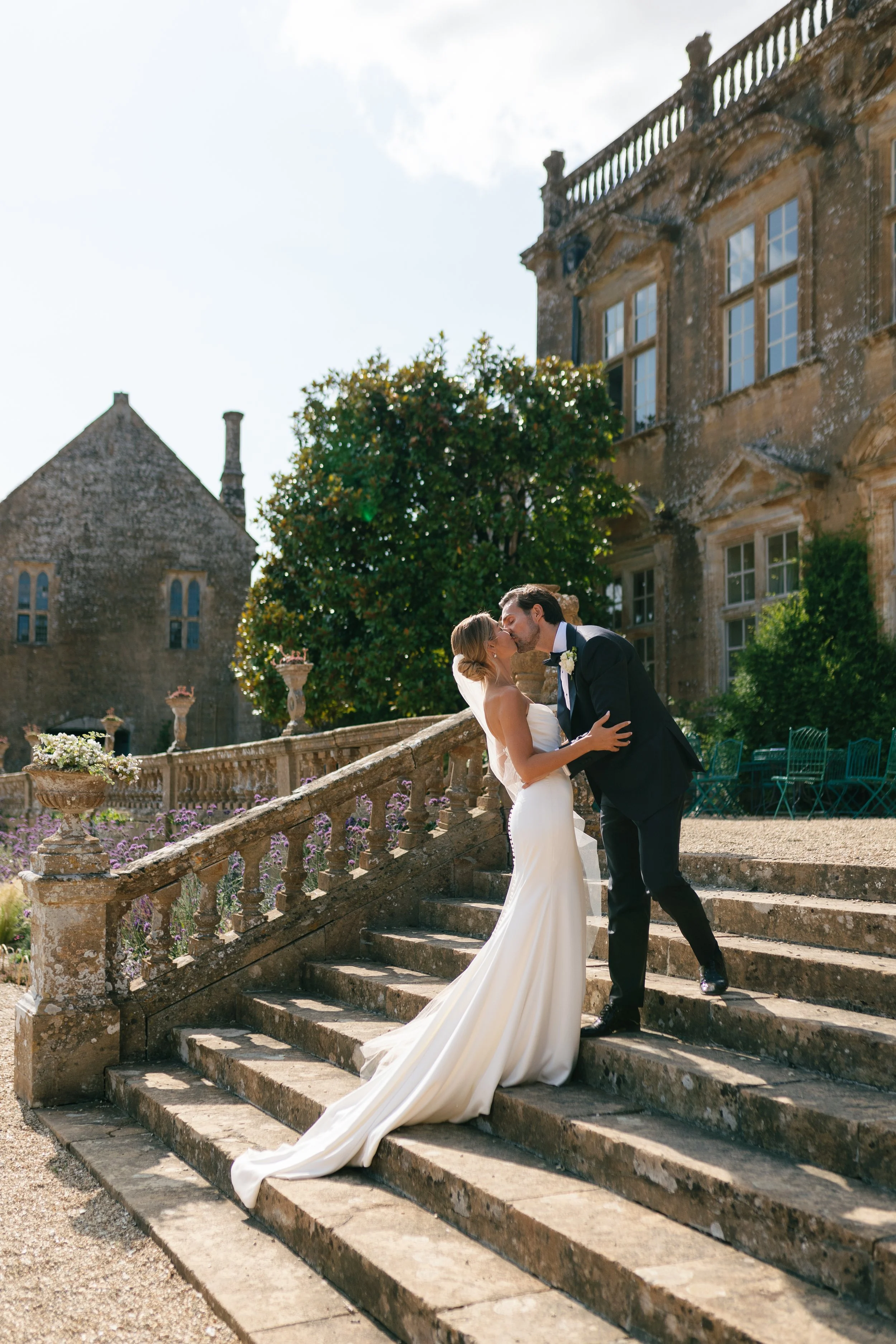 A bride and groom kissing on stone steps outside a historic building with large windows and a lush tree.