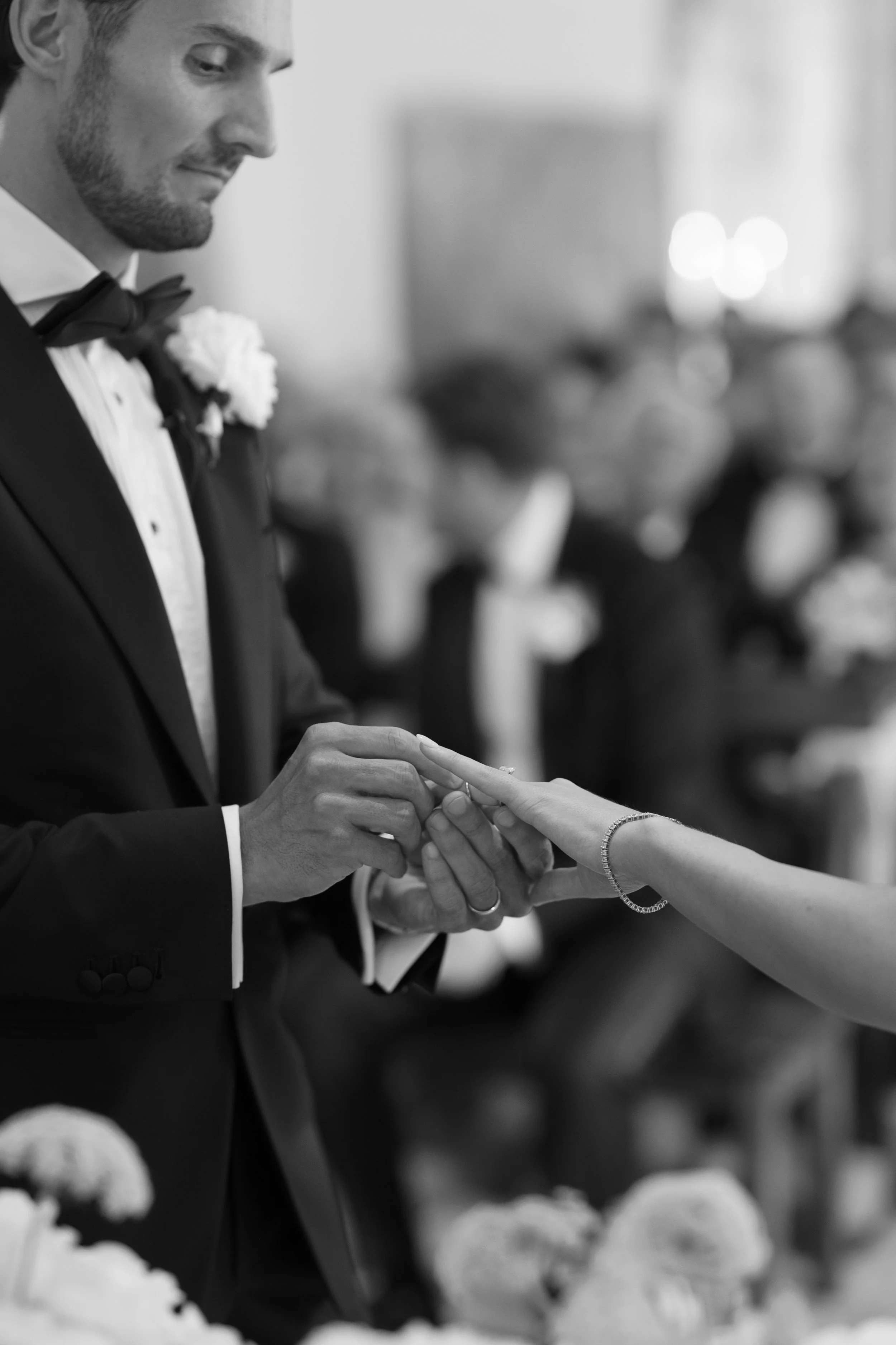 A groom places a wedding ring on the bride's finger during a wedding ceremony, with guests seated in the background.