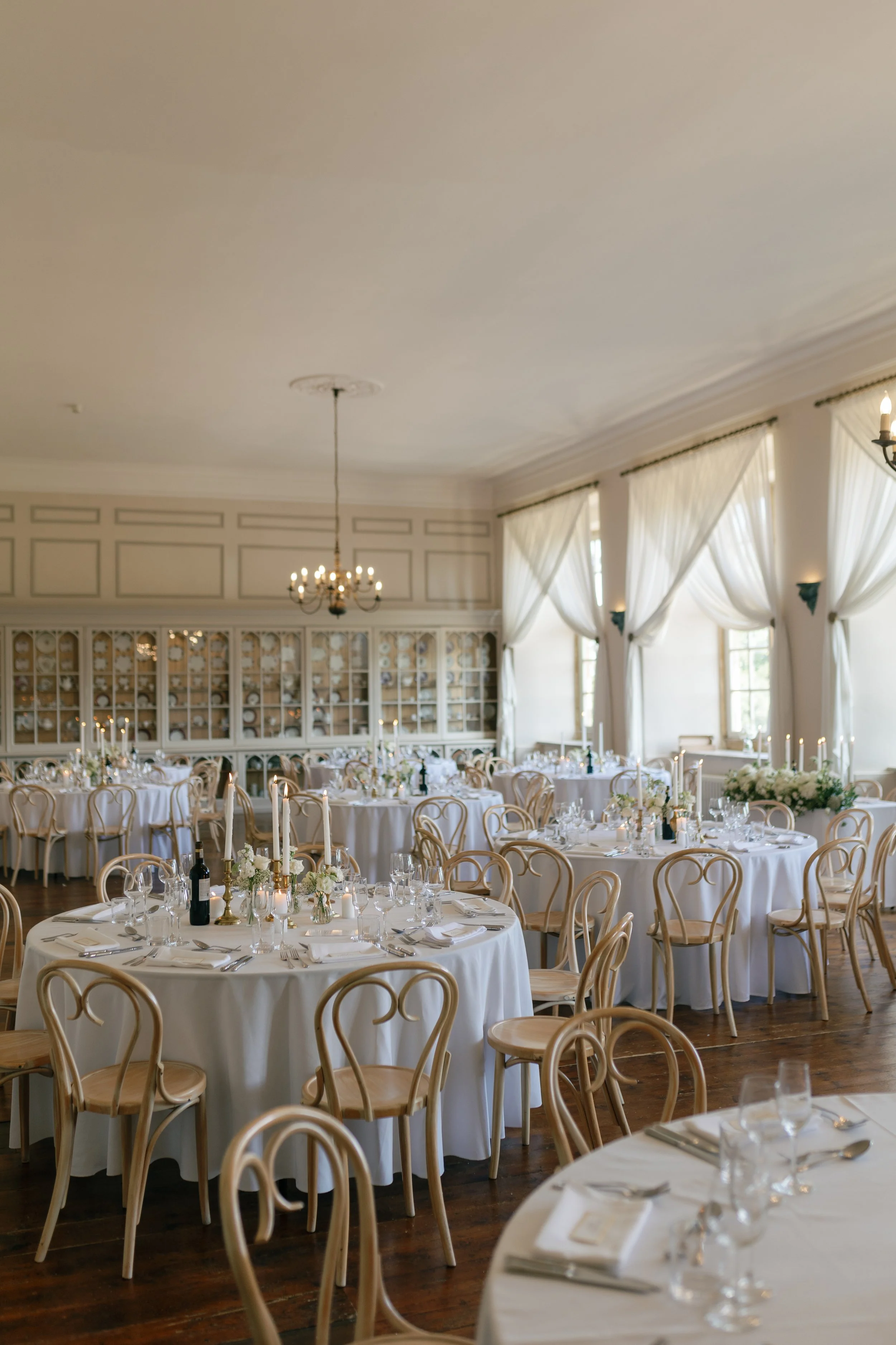 Elegant banquet hall with round tables draped in white tablecloths, set for a formal event, decorated with floral centerpieces, candles, and glassware. Large windows with white curtains allow natural light, and a chandelier hangs from the ceiling.