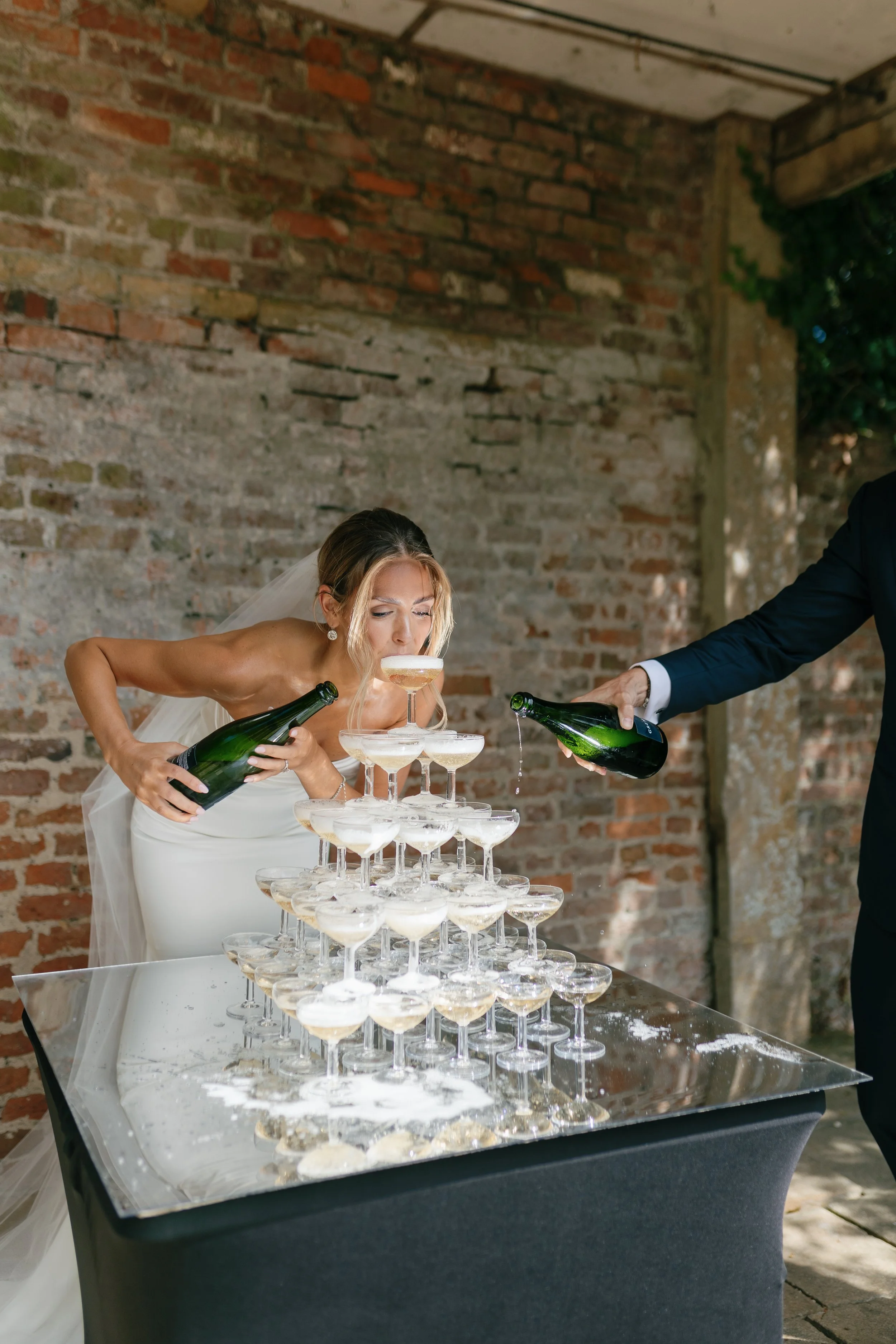 Bride in wedding dress drinks champagne from a pyramid of glasses at a wedding reception, with a person pouring more champagne.