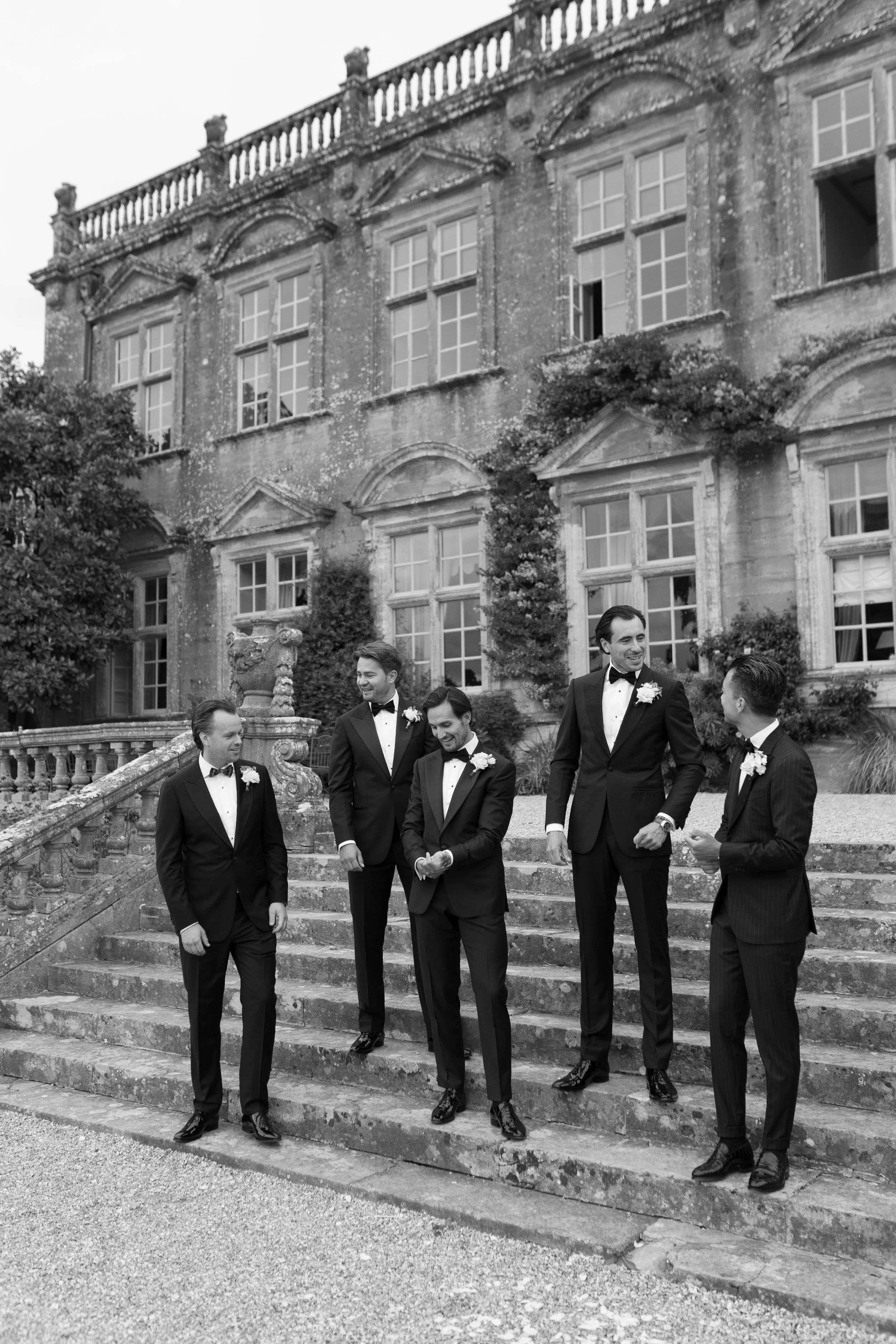 A group of five men in tuxedos standing on stone steps outside a large historic building, engaging in conversation and smiling.