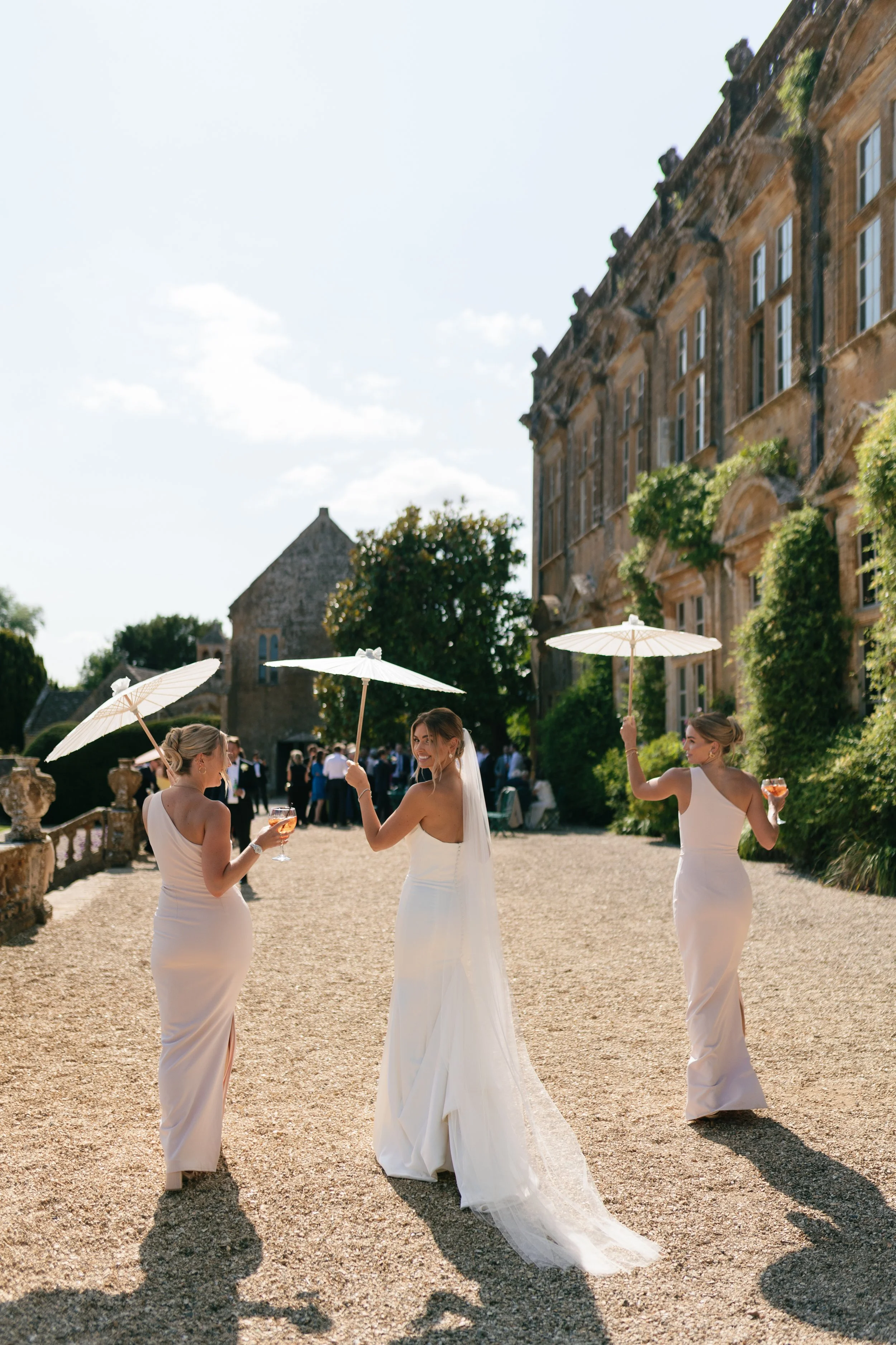 Bride and two bridesmaids holding umbrellas and glasses of wine at outdoor wedding in sunlight