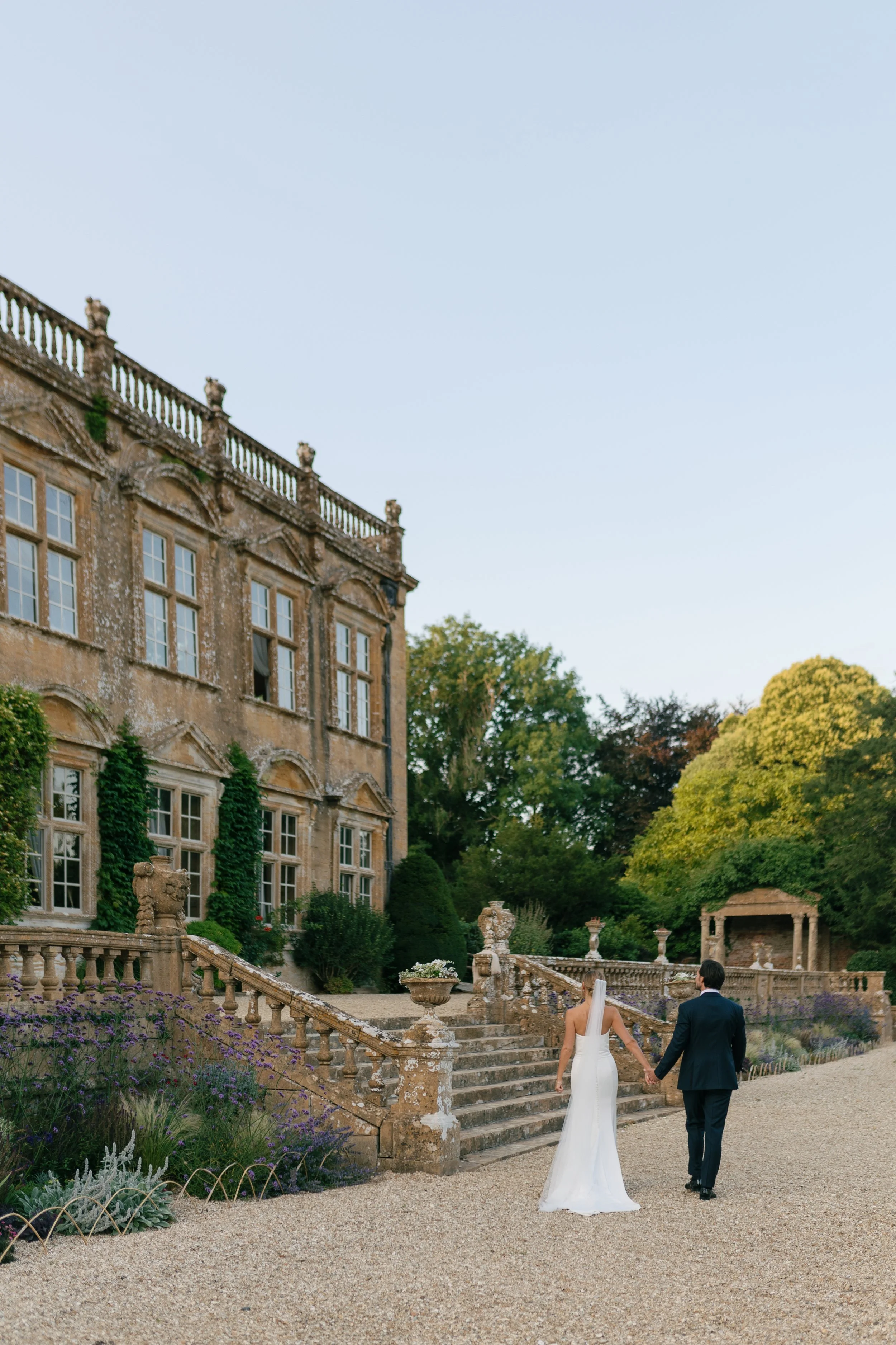 A bride and groom walking hand in hand outside a historic estate with stone stairs and lush greenery.