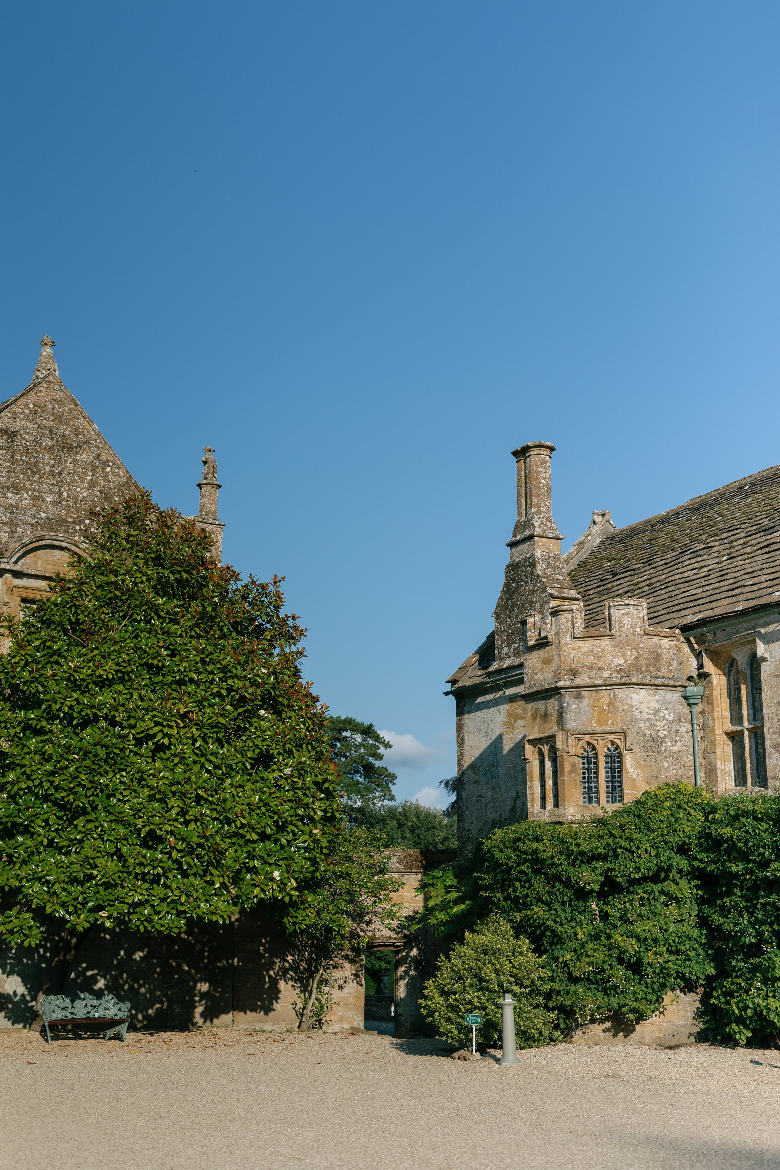 Historic stone building with a chimney and arched windows, surrounded by greenery and a gravel courtyard under a clear blue sky.