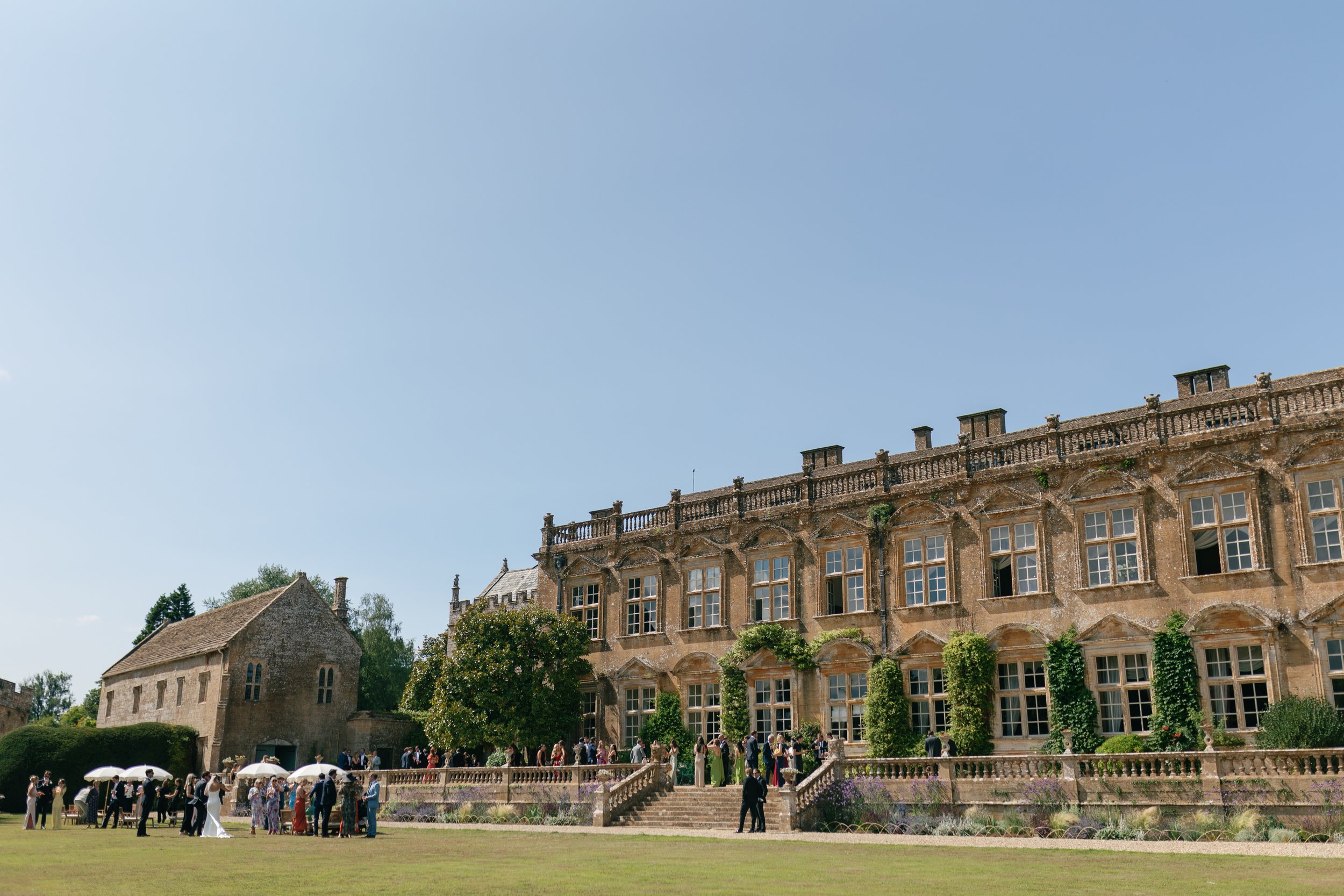 A large historic mansion with multiple windows and a stone staircase, surrounded by guests at an outdoor event with umbrellas on a sunny day.