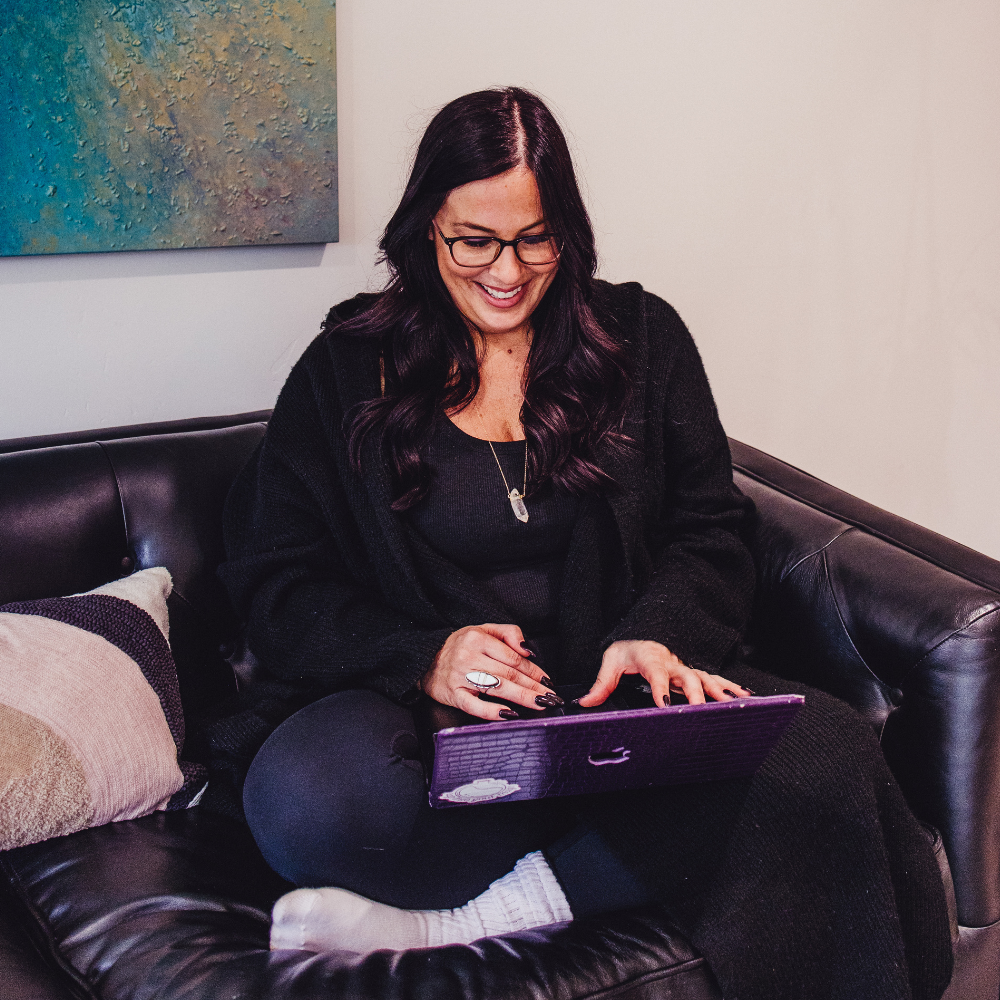 woman smiling, sitting on a leather couch, typing on her laptop