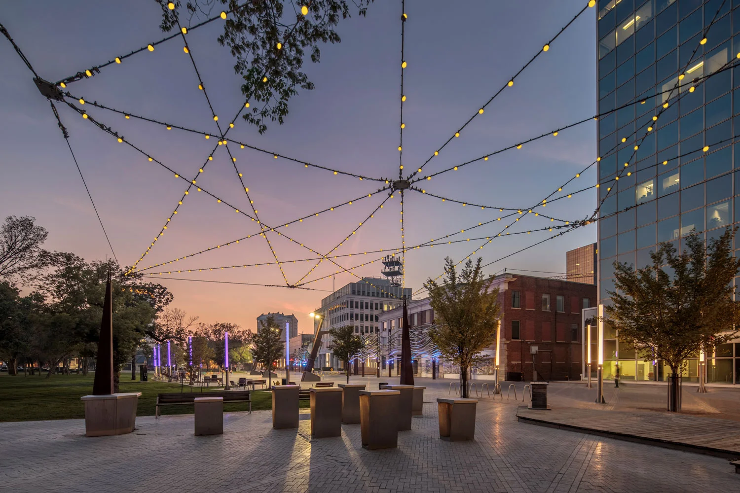 ….This plaza twinkles at night with multiple overhead LED lights. The space is a transitional connection between Victoria Park and the Scarth Street Mall, a well-used pedestrian street north of the plaza. Custom-made stainless steel tables were designed for casual lunches. ..En soirée, la place publique scintille grâce à de multiples dispositifs d’éclairage aérien DEL. Le lieu sert d’espace de transition reliant Victoria Park au Scarth Street Mall, une rue piétonne au fort achalandage, au nord de la place publique. Des tables en acier inoxydable furent conçues sur mesure afin d’accommoder les pauses repas. ….